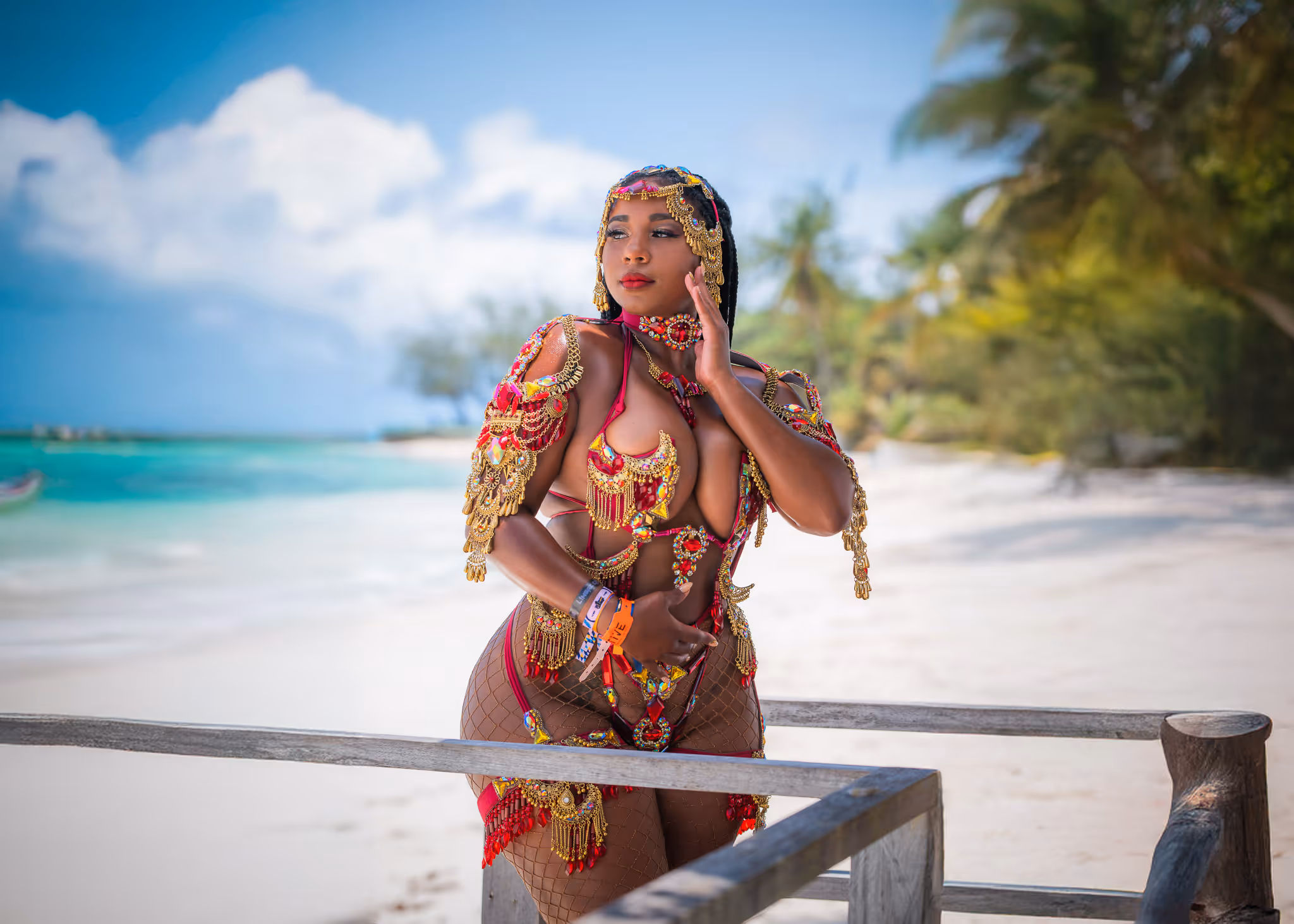 Woman in ornate red and gold carnival costume posing on a wooden railing by a tropical beach.