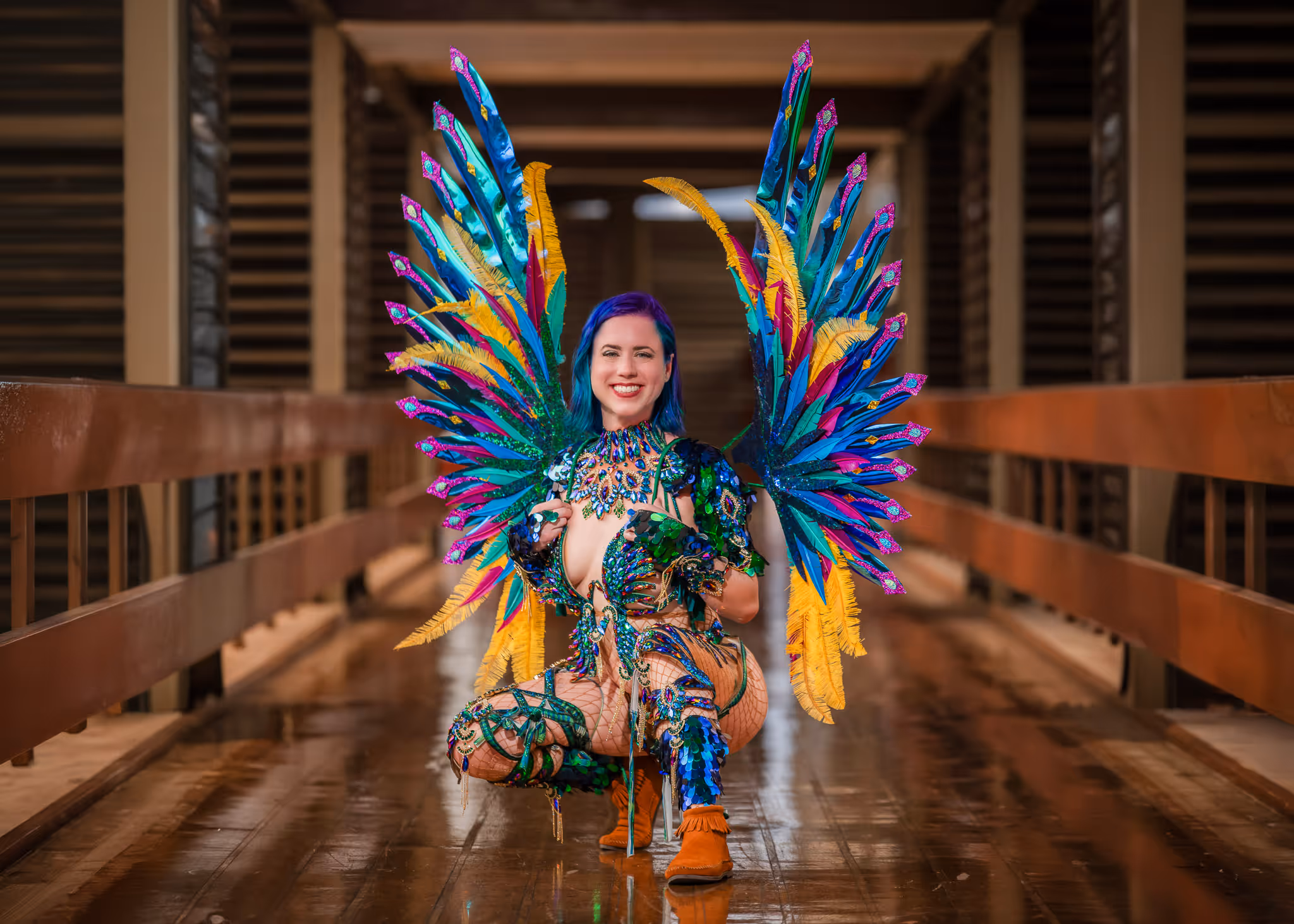 Smiling person with blue hair wearing colorful, elaborate carnival wings and costume squatting on a wooden bridge.