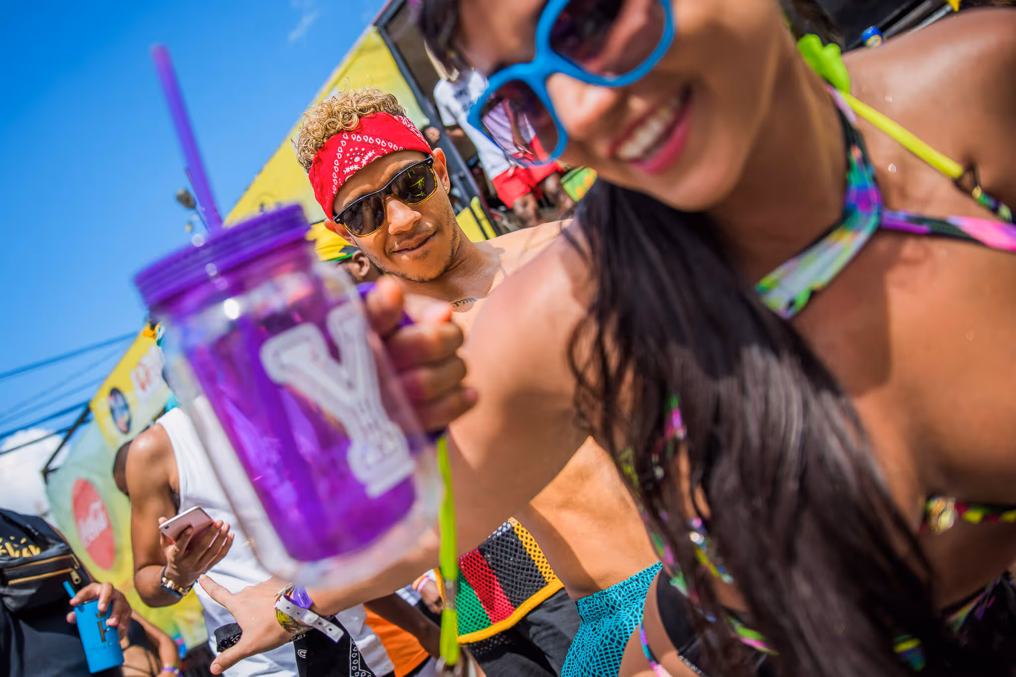 Smiling man wearing sunglasses and red bandana and a woman in colorful sunglasses holding a purple cup at outdoor festival.