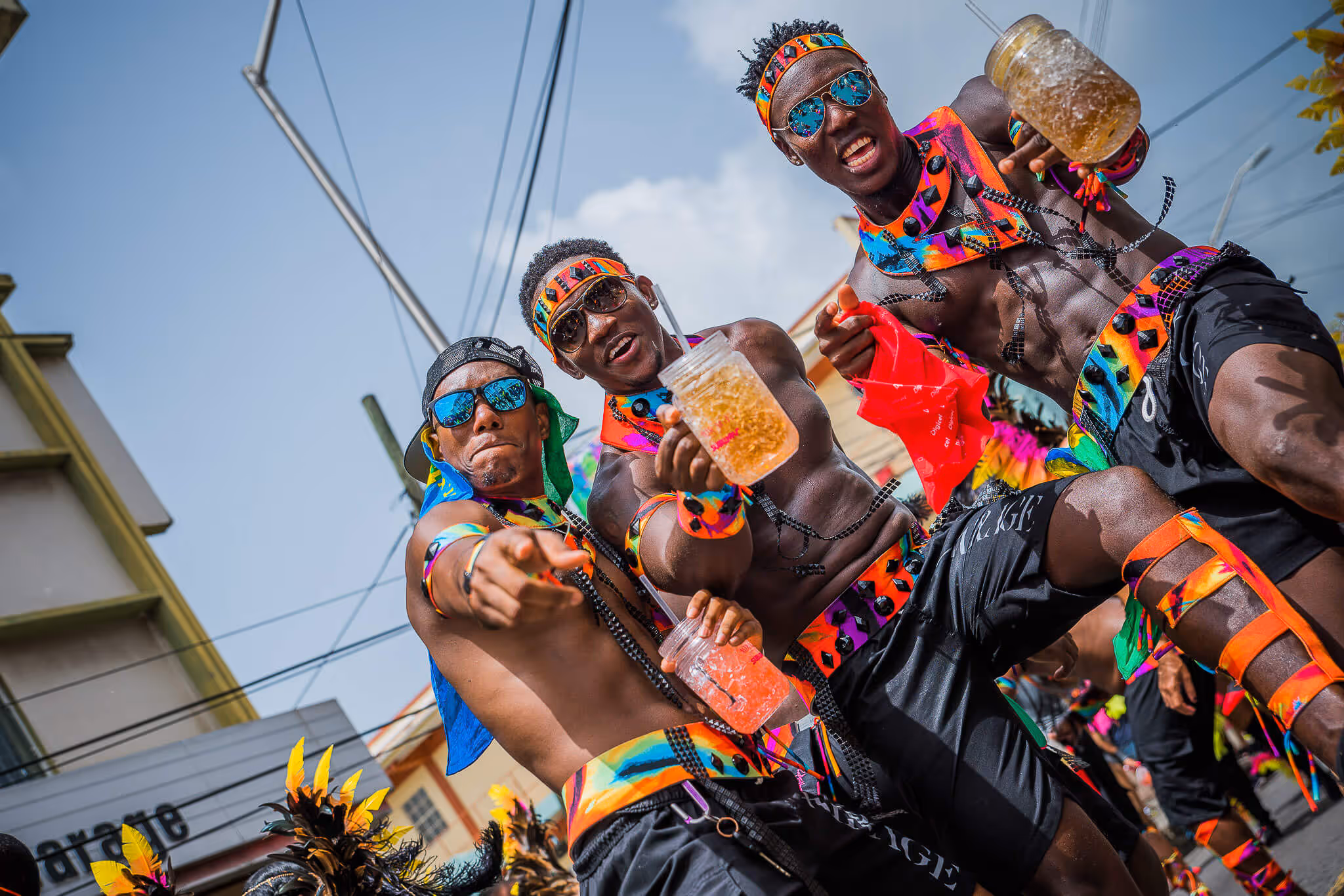 Three men in vibrant carnival costumes and sunglasses enjoying a street festival, holding drinks and pointing towards the camera.