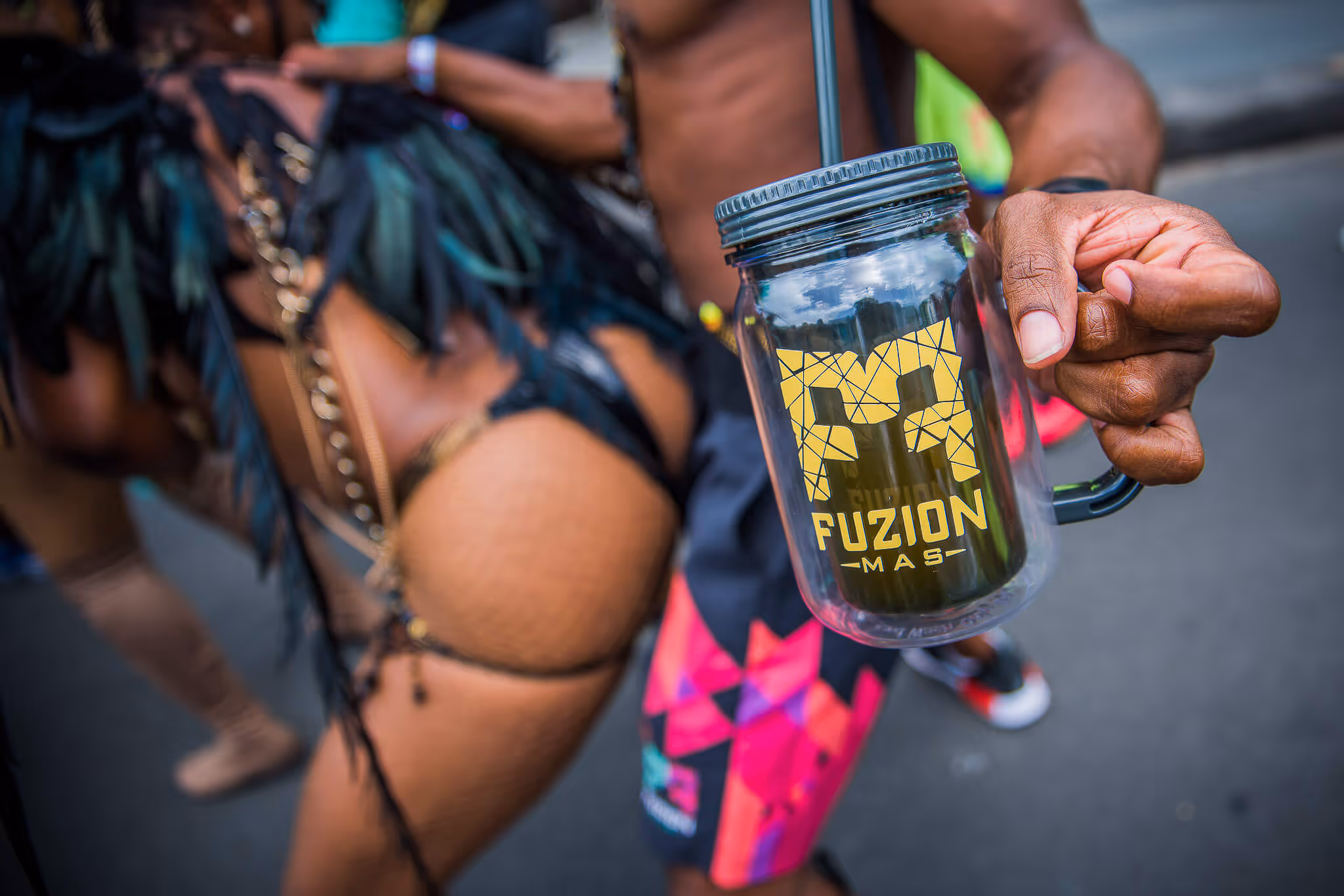 Close-up of a hand holding a clear plastic mason jar labeled 'Fuzion Mas' with blurred carnival dancers in colorful costumes in the background.