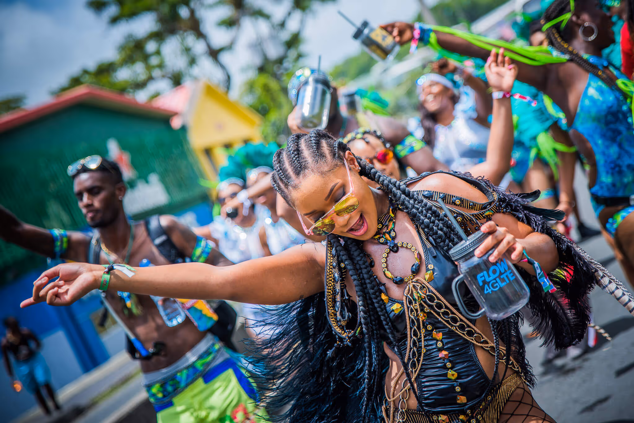 Woman with braided hair dancing energetically in colorful carnival attire, holding a cup that reads 'FLOW 4GLTE', surrounded by other festive dancers.