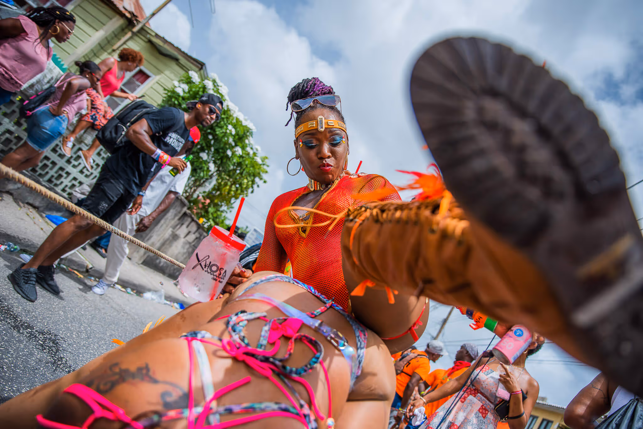 Woman in vibrant carnival costume dancing with one leg raised, holding a cup, surrounded by onlookers on a street.