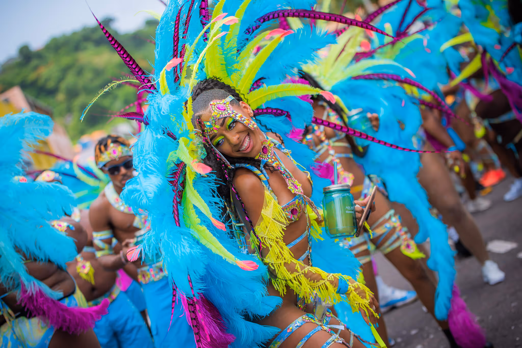 Smiling woman wearing bright carnival costume with blue and yellow feathers holding a green drink in a mason jar during a street parade.