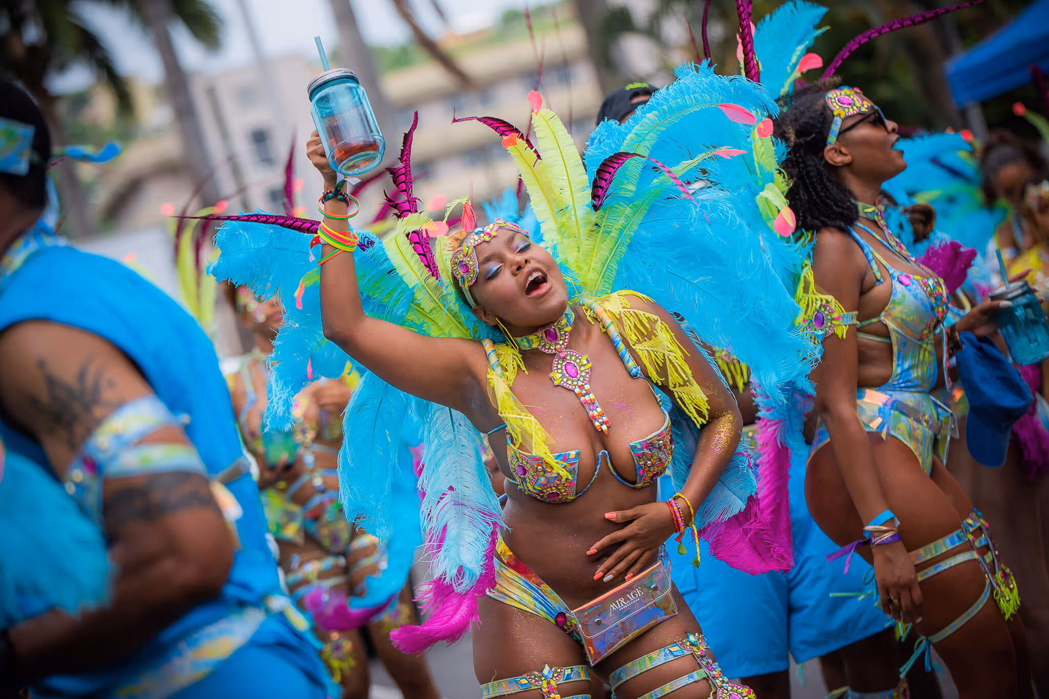 Woman dancing joyfully in colorful carnival costume with blue and yellow feathers, holding a drink jar.