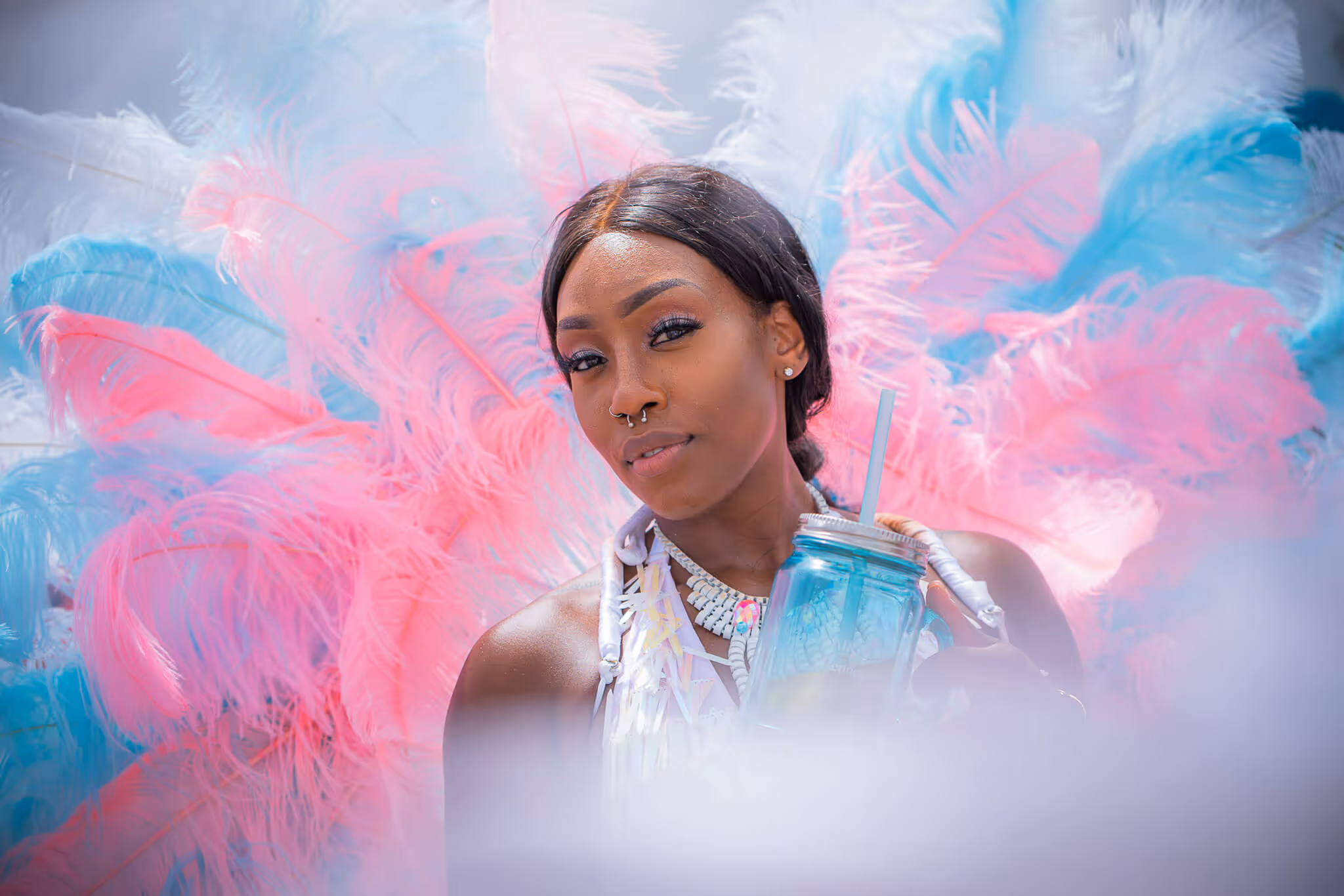 Woman with septum piercing and black hair posing with a blue feathered costume and holding a blue mason jar with a straw.