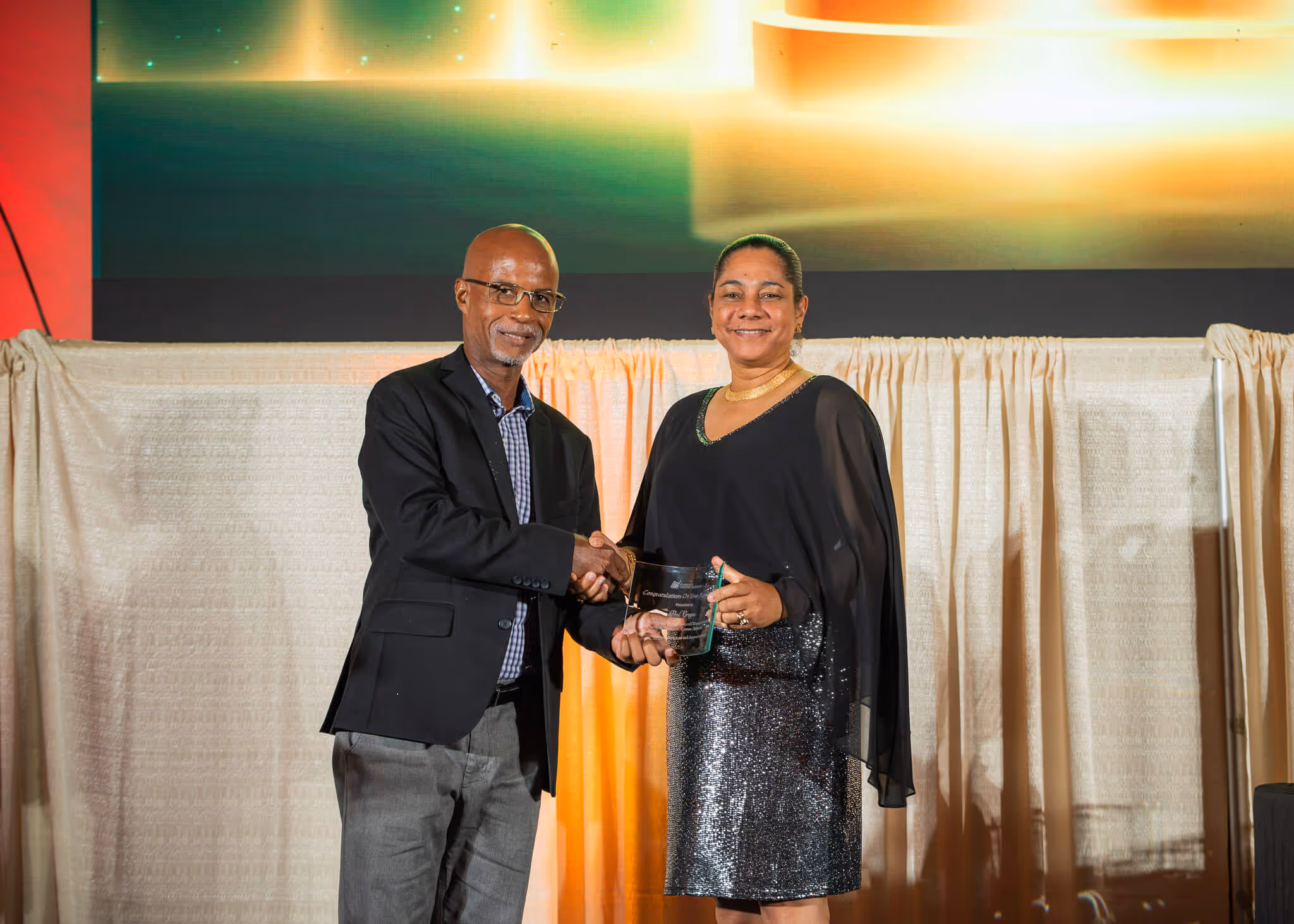 Man in a suit shaking hands with a woman in a black dress holding an award plaque on stage.