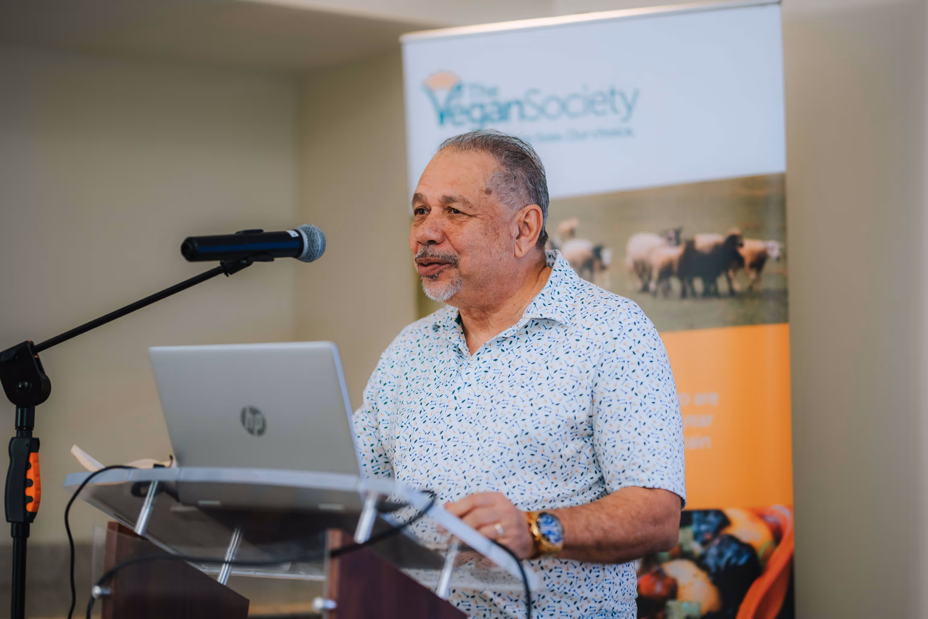 Man speaking at a microphone behind a podium with a laptop, standing in front of a banner for The Vegan Society.