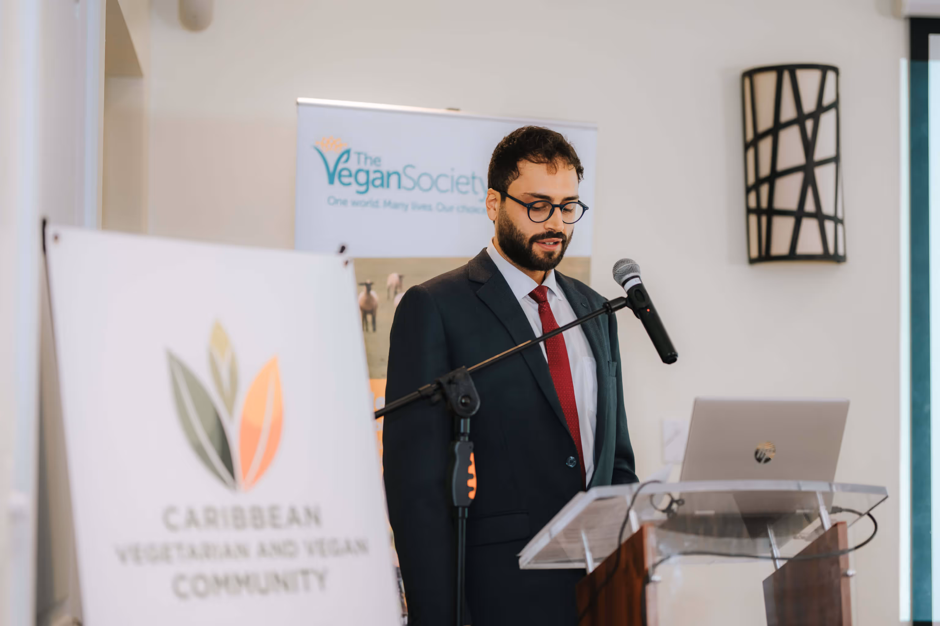 Man in a suit with glasses speaking at a podium with a microphone and laptop, with banners for The Vegan Society and Caribbean Vegetarian and Vegan Community.