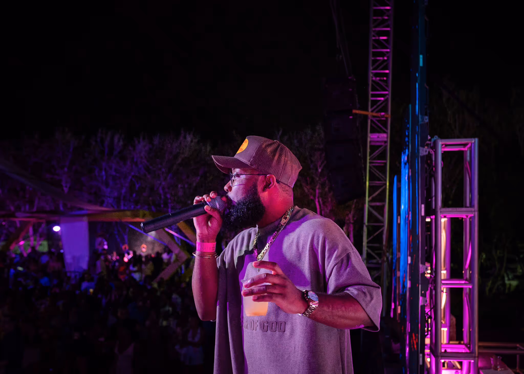 Bearded man wearing a cap and glasses rapping into a microphone on stage at night, holding a drink in his other hand.