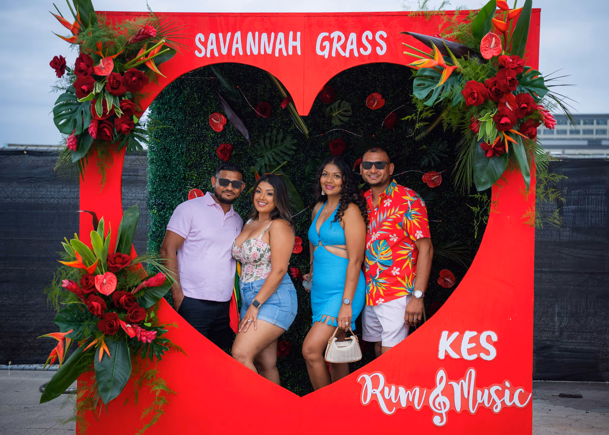 Four people posing inside a large red heart-shaped frame decorated with tropical flowers, with text 'Savannah Grass,' 'KES,' and 'Rum & Music'.