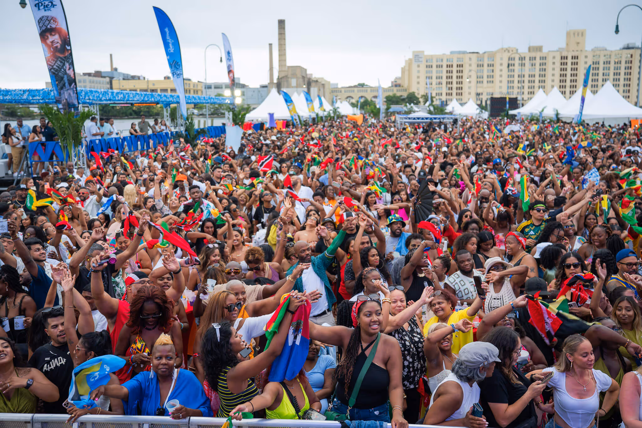 Large lively crowd at an outdoor festival waving colorful flags with buildings and white tents in the background.
