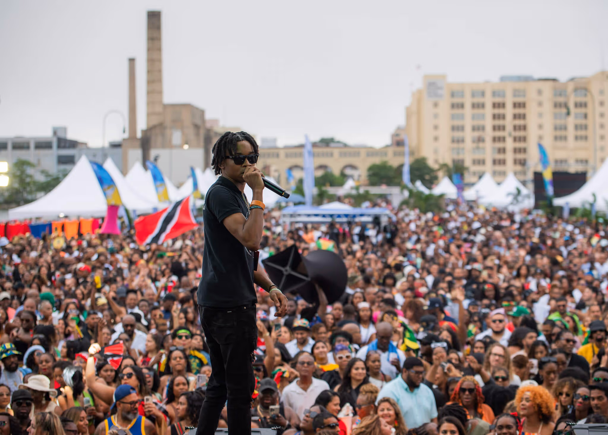Male performer wearing sunglasses and black clothes singing into a microphone on stage in front of a large outdoor crowd.