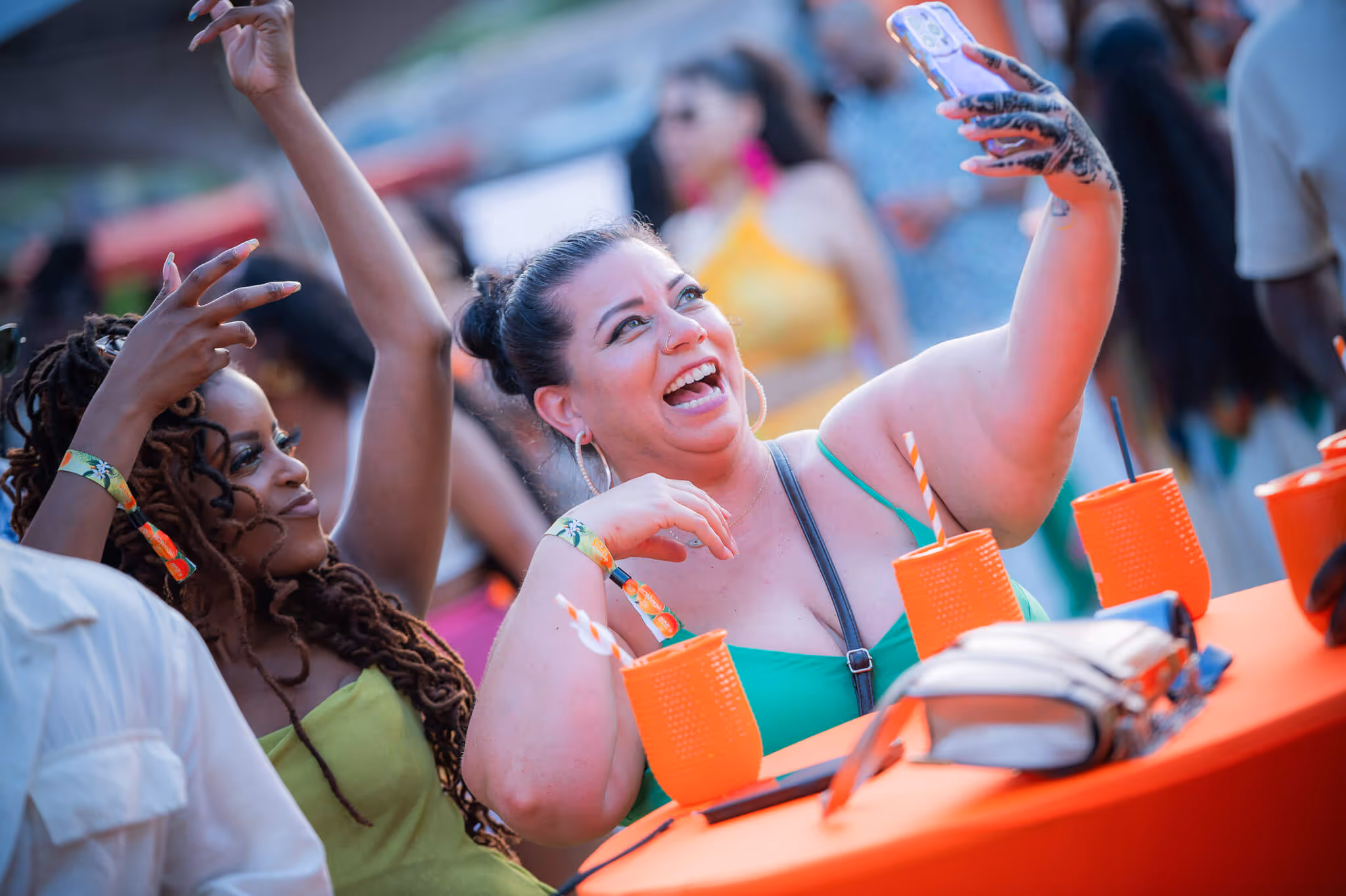 Two women at an outdoor event, one in green taking a selfie with a decorated hand and the other posing with raised arms, surrounded by orange cups.