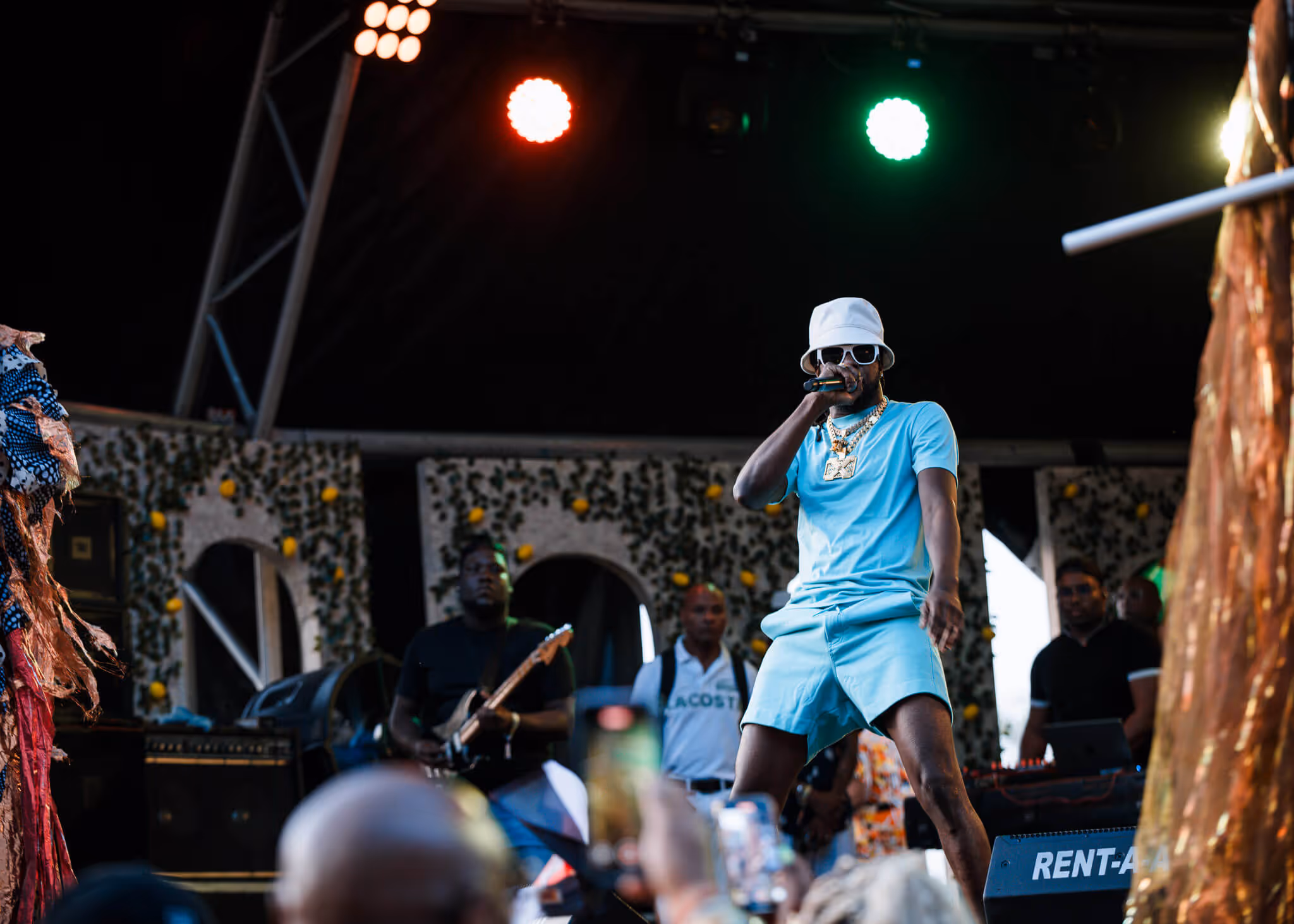 Male performer in light blue outfit and white bucket hat singing on stage with musicians and colorful stage lights behind.