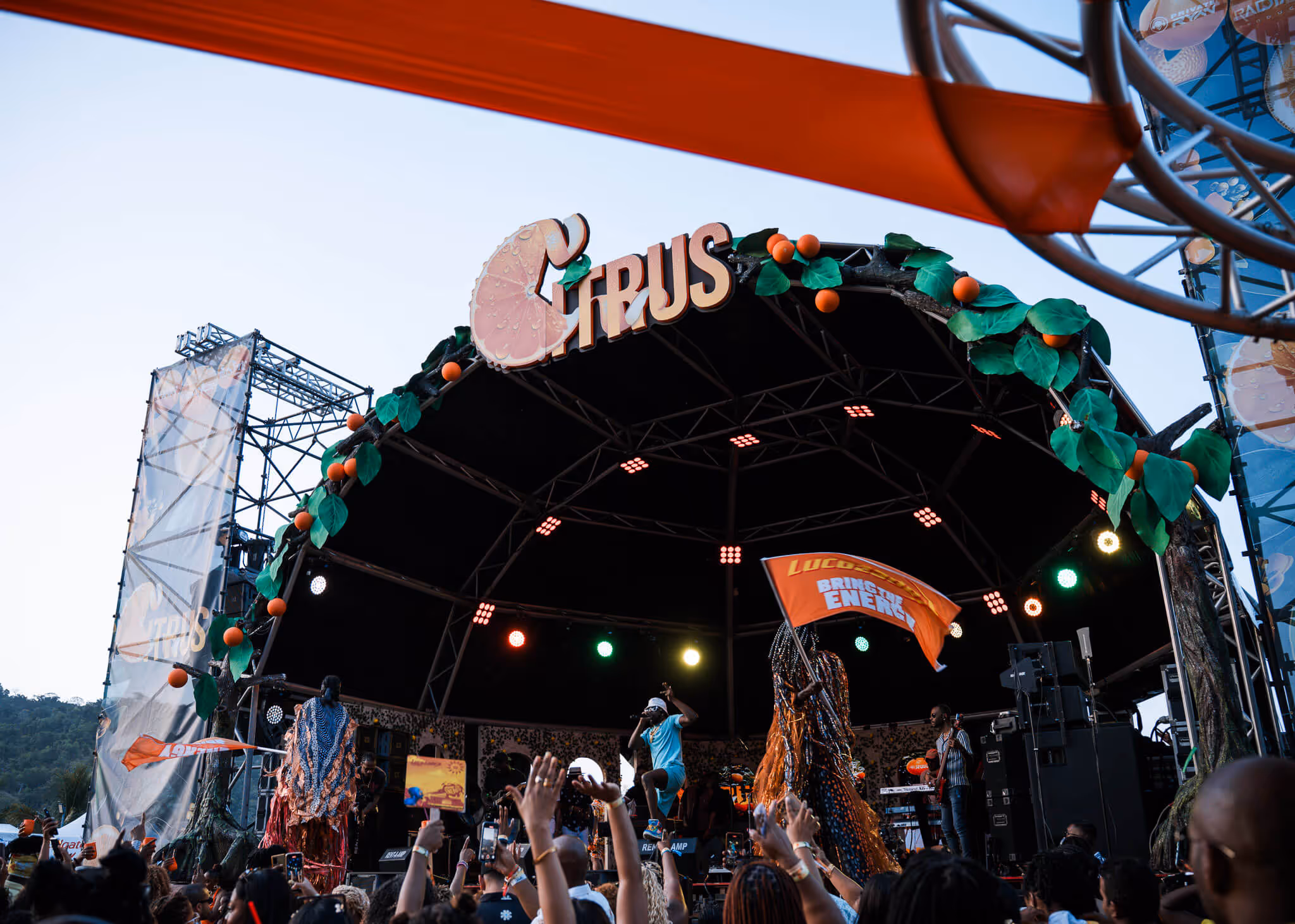 Outdoor concert stage decorated with green leaves and orange fruits, performers on stage, and audience raising hands.