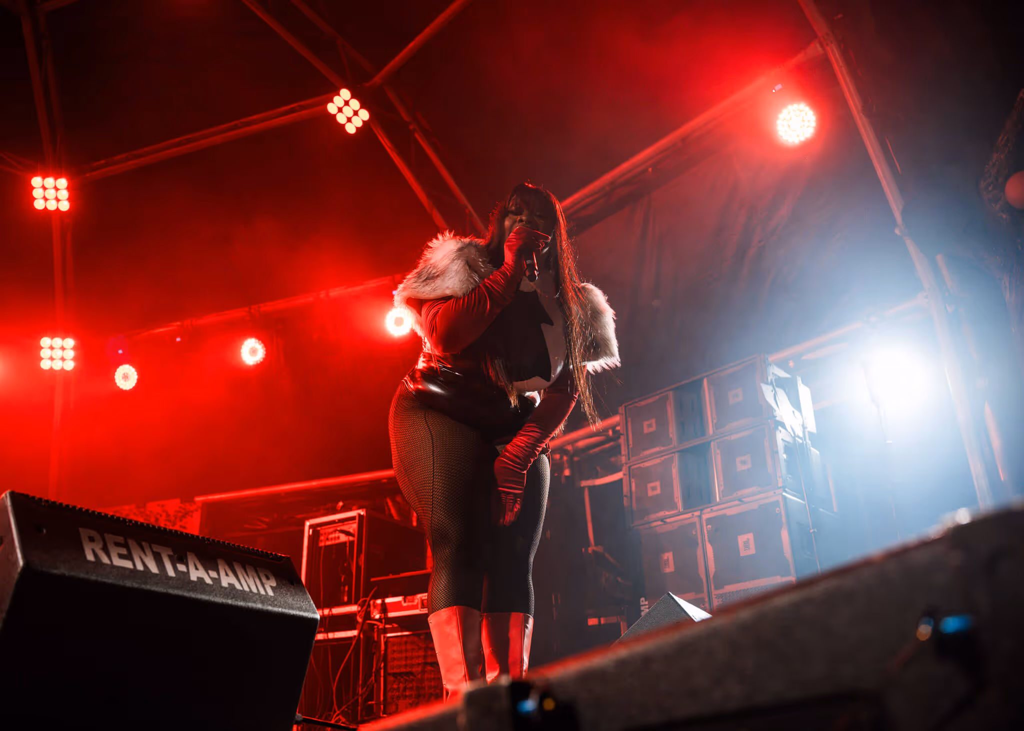 Female singer wearing red gloves, boots, fishnet leggings, and a fur-shouldered outfit performing on stage with red and white lighting.