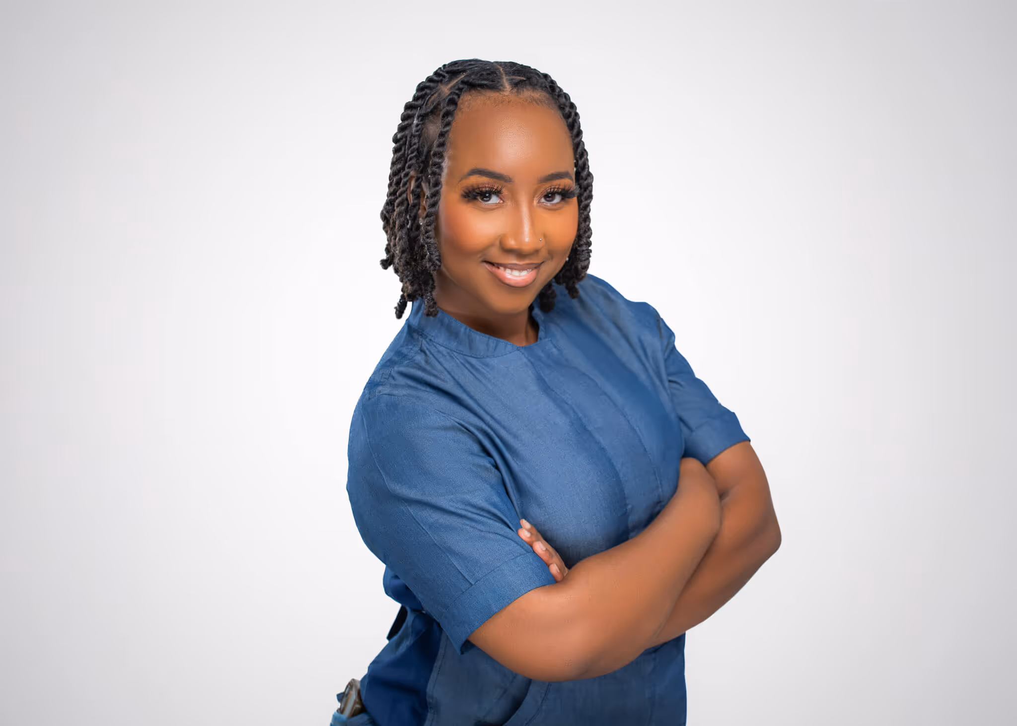 Smiling woman with braided hair wearing a blue shirt and crossing her arms against a light gray background.