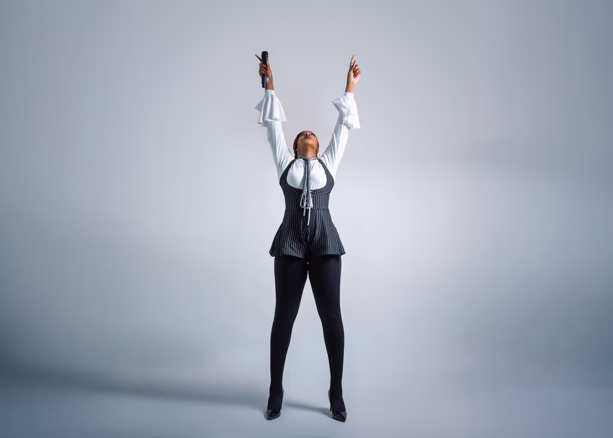 Woman standing with arms raised, holding a microphone, dressed in a white blouse and black pinstripe outfit against a gray background.
