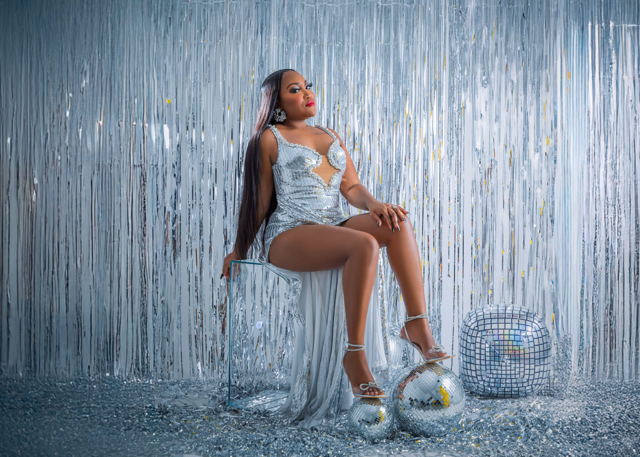 Woman in a sparkly silver dress sitting on a transparent chair with disco balls and silver streamers in the background.