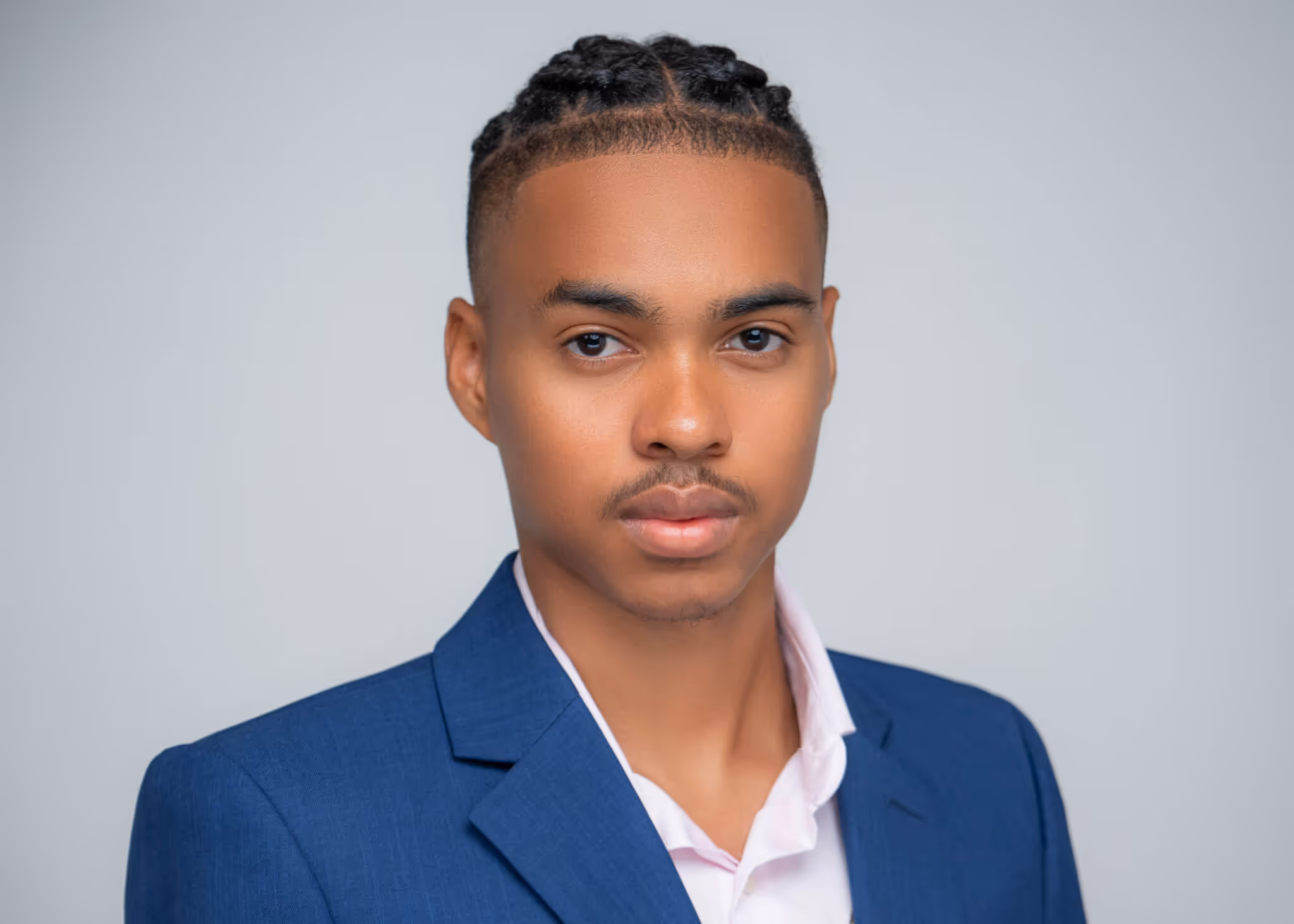Young man with braided hair wearing a blue blazer and white shirt against a gray background.