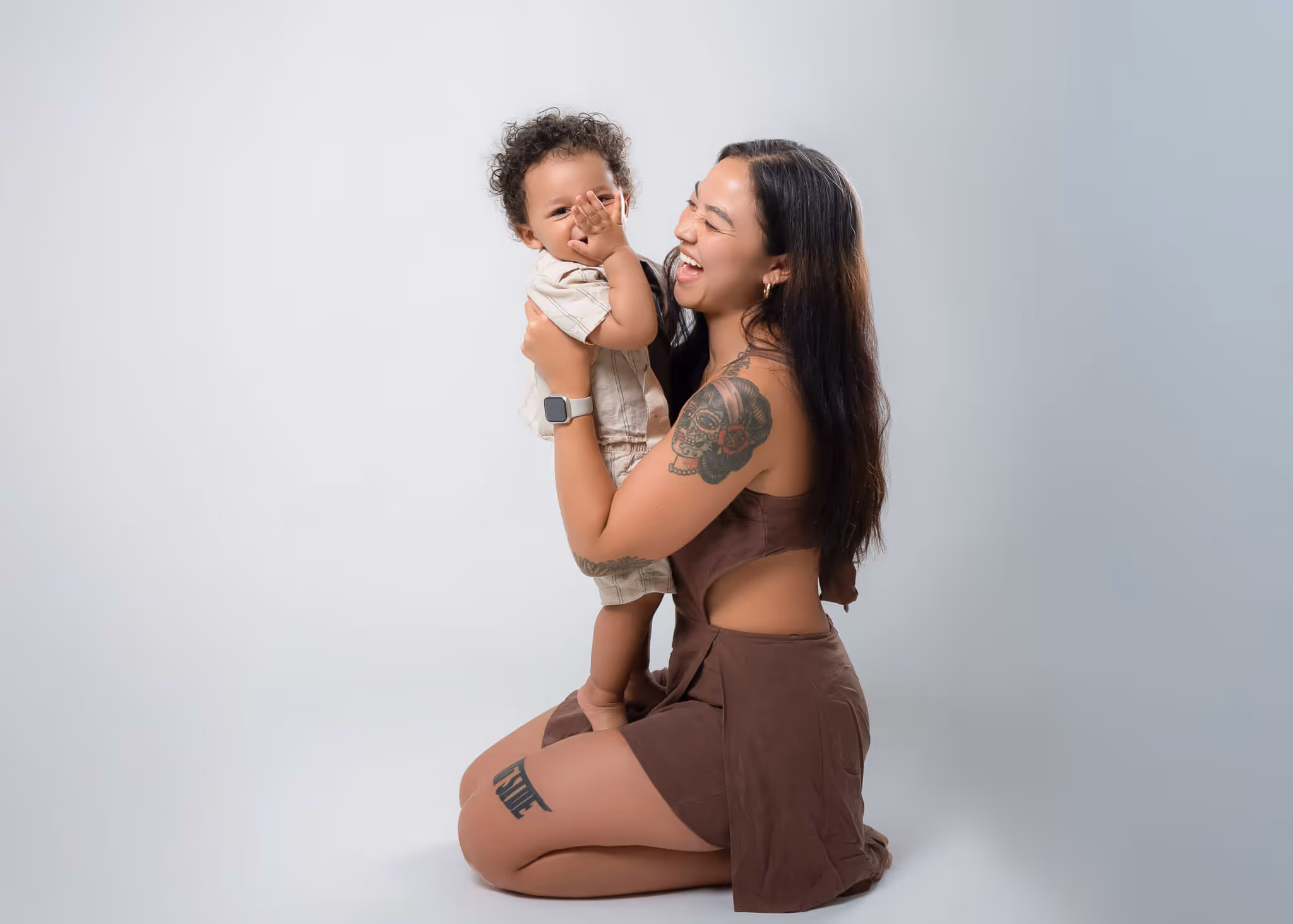 Woman with tattoos kneeling and playfully holding a smiling toddler in beige clothes against a plain light background.