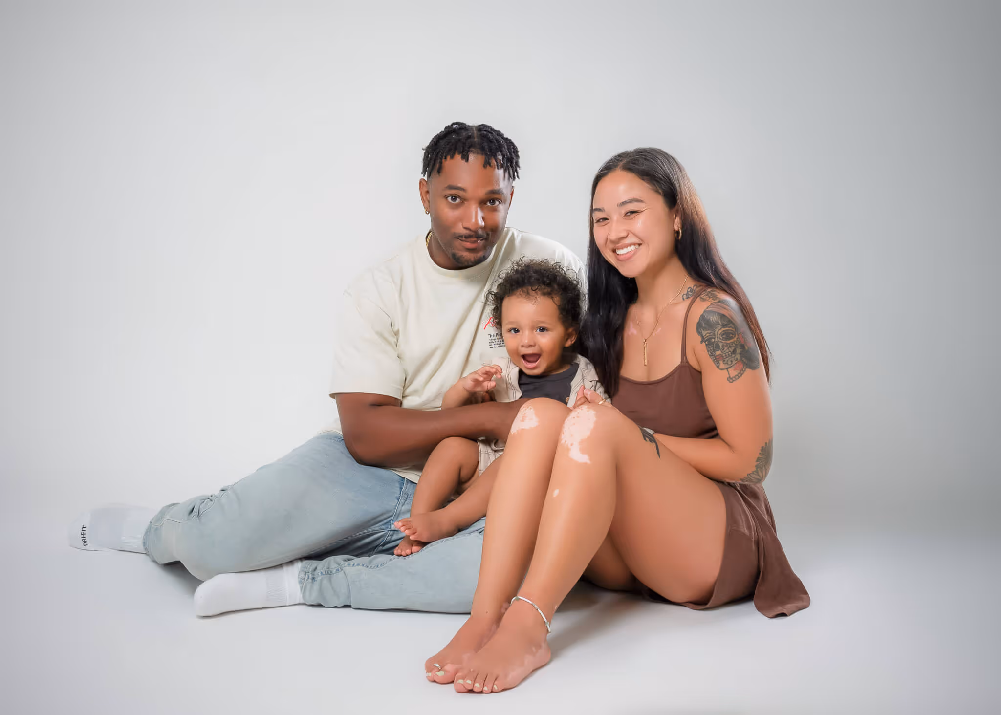 Smiling family of three seated on the floor, including a man, woman with tattoos, and a young child.
