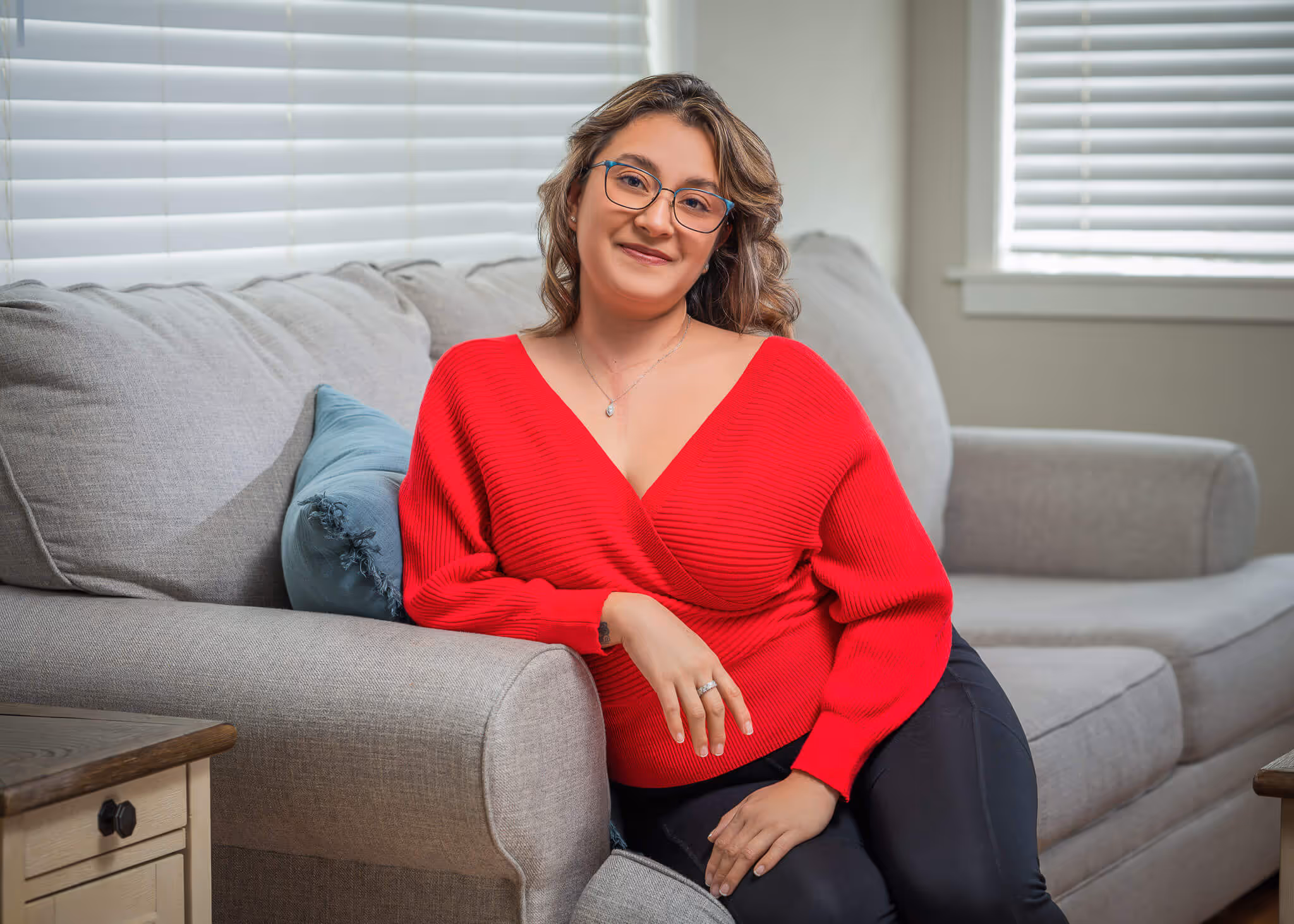 Woman with glasses wearing a red sweater sitting on a gray couch in a living room.