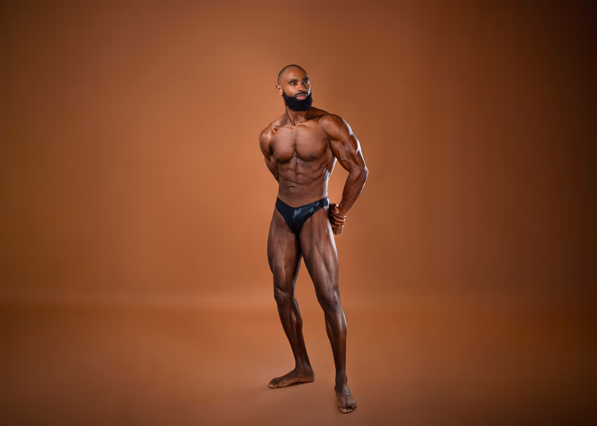 Muscular man with a beard posing in black briefs against a brown background.