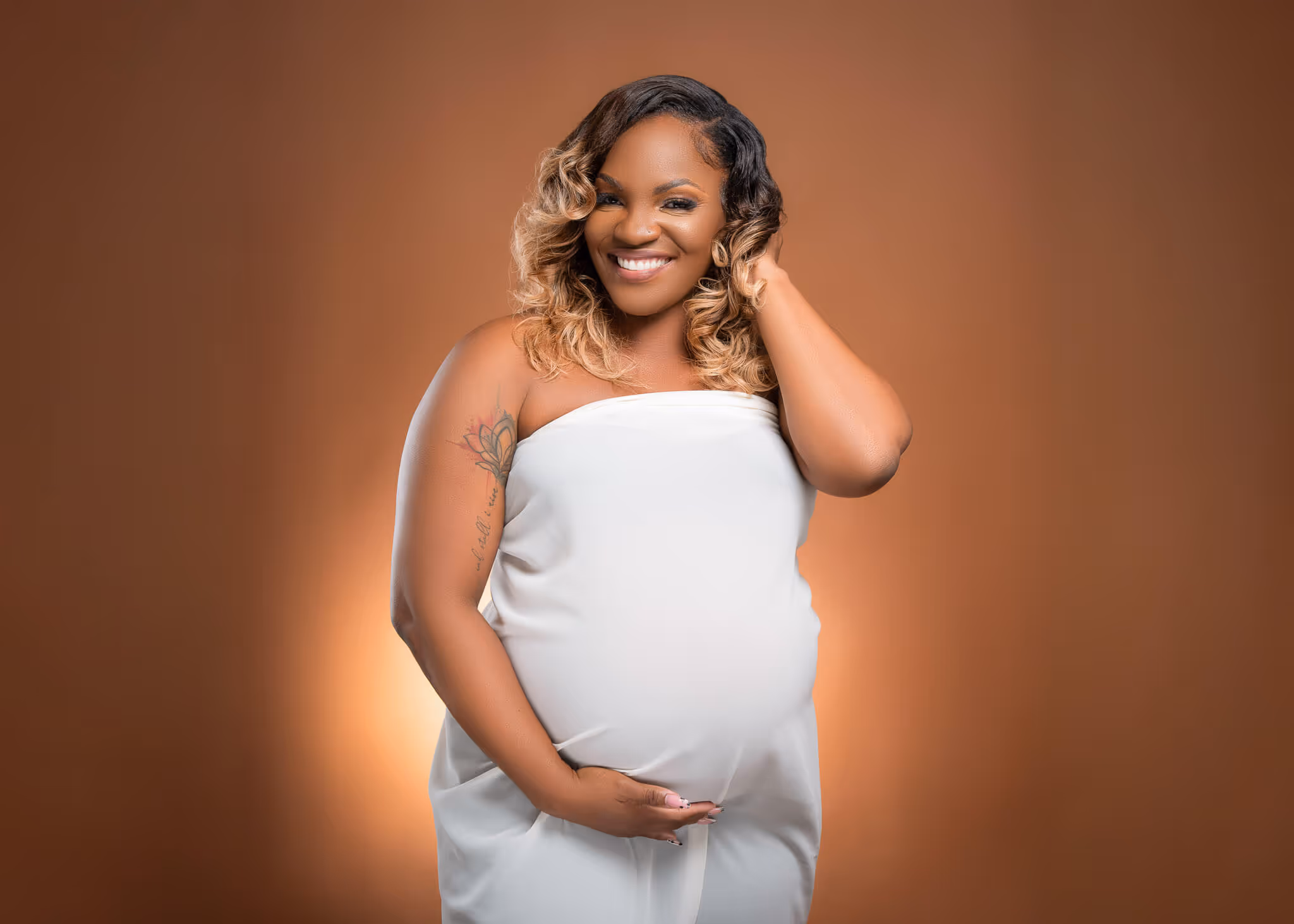 Smiling pregnant woman in white dress holding her belly against a brown background.