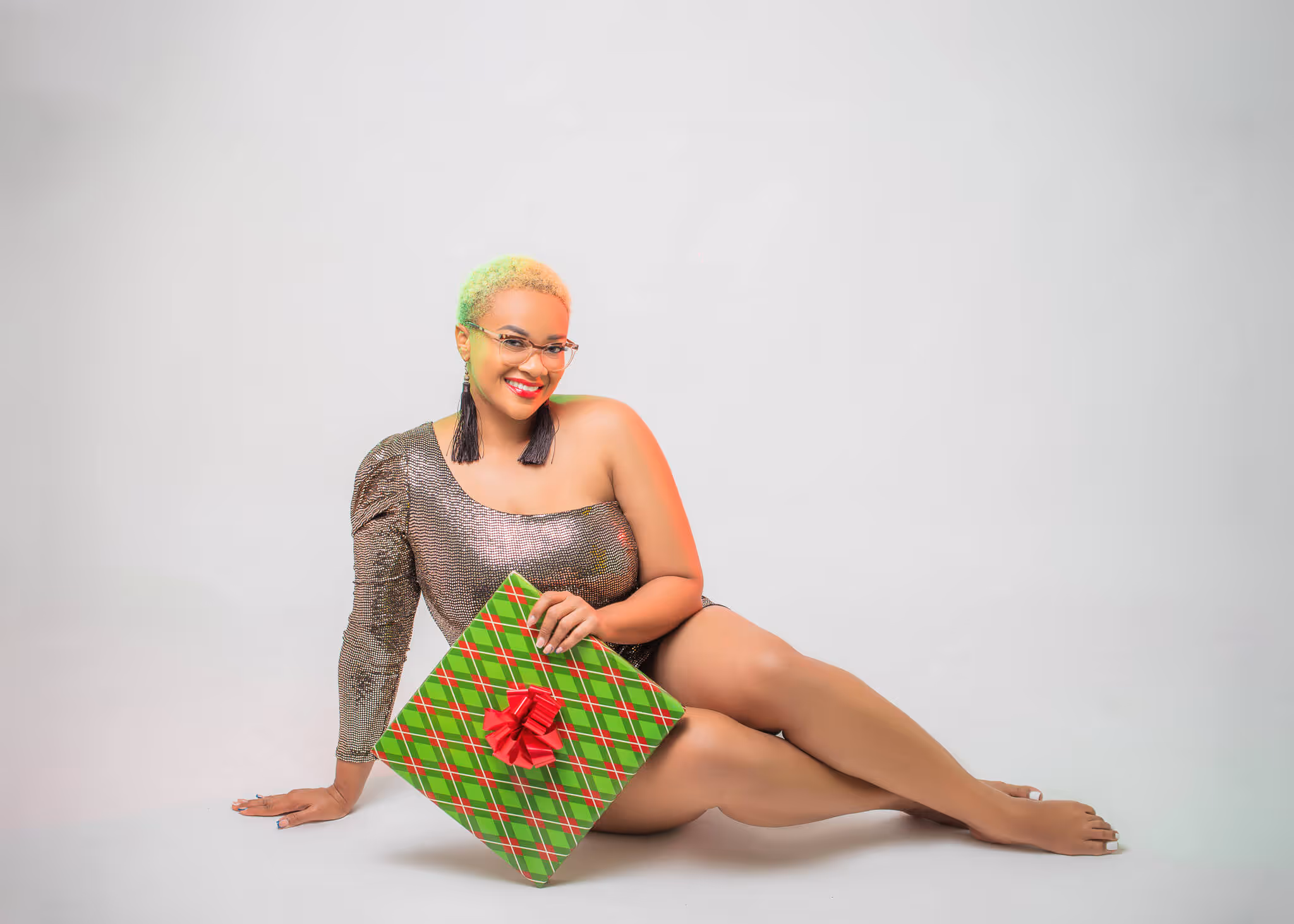 Smiling woman with short blonde hair and glasses sits on the floor holding a green and red plaid wrapped gift with a red bow.