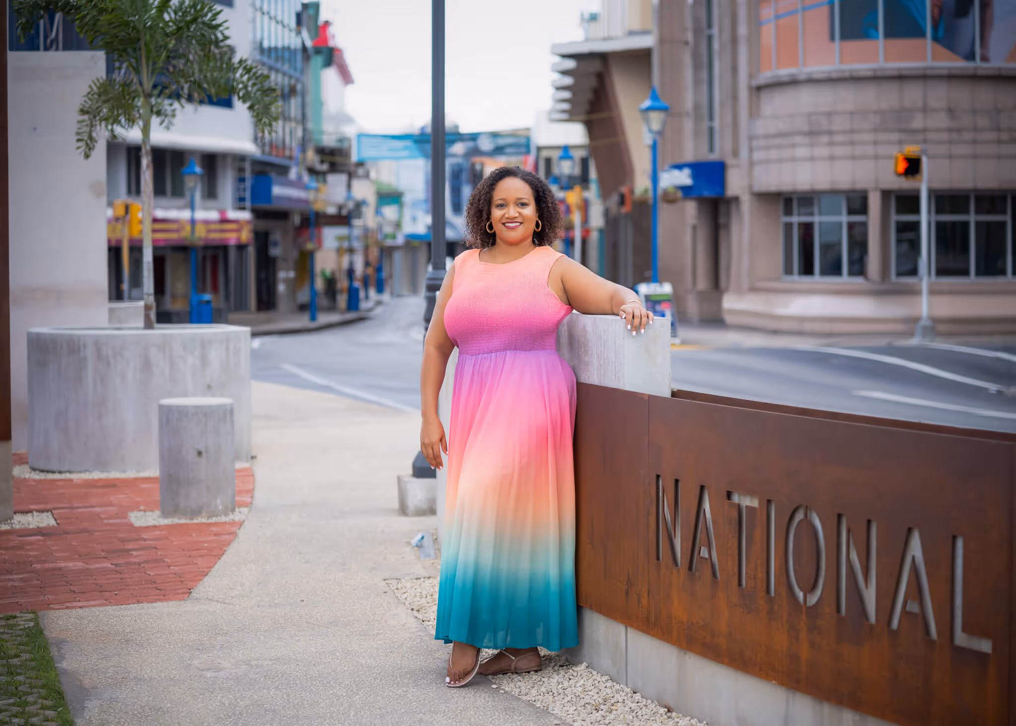 Woman in a colorful ombre dress leaning on a large sign with the word 'NATIONAL' on a city street.