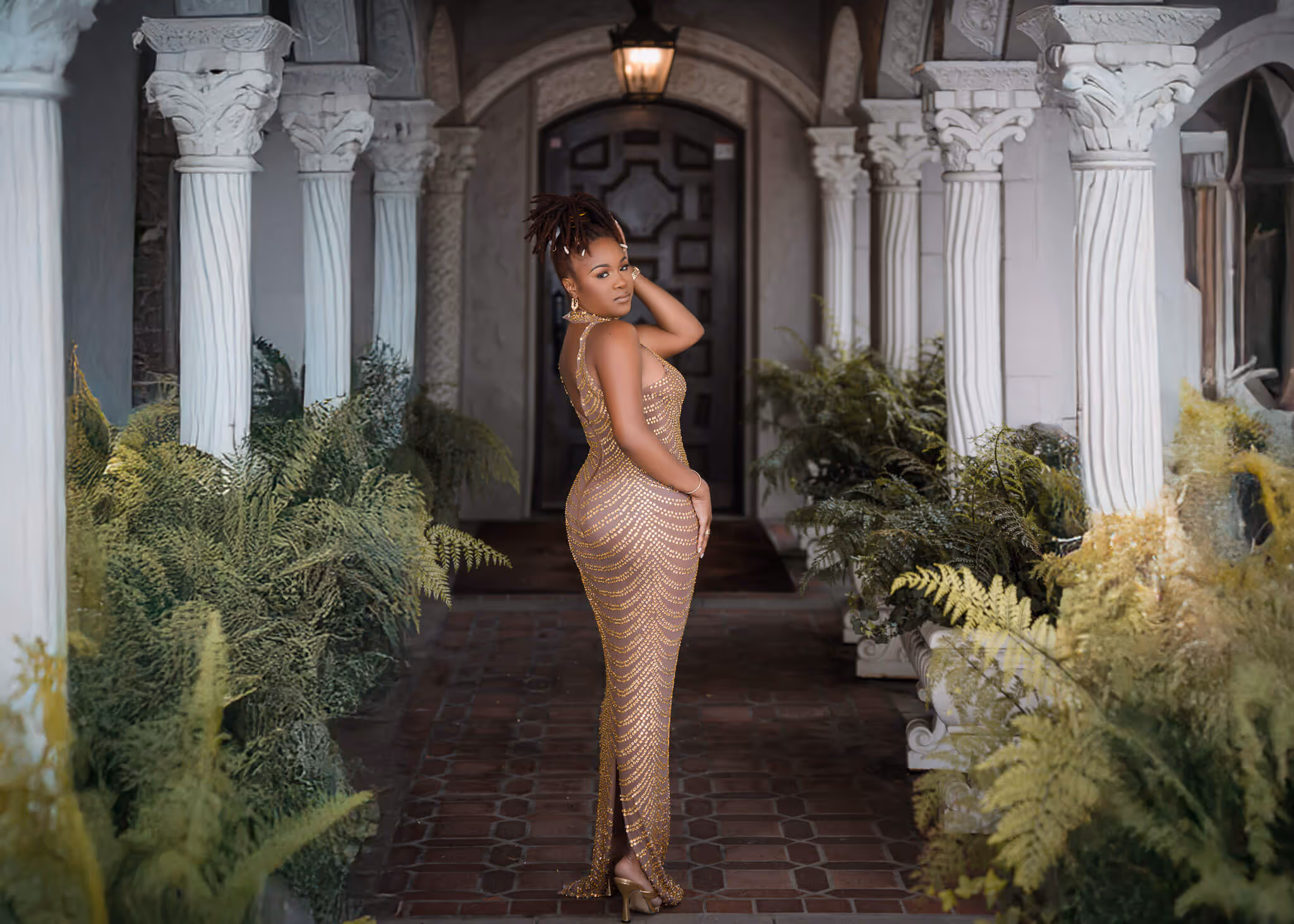Woman in a fitted gold dress posing in a corridor with white columns and green plants.