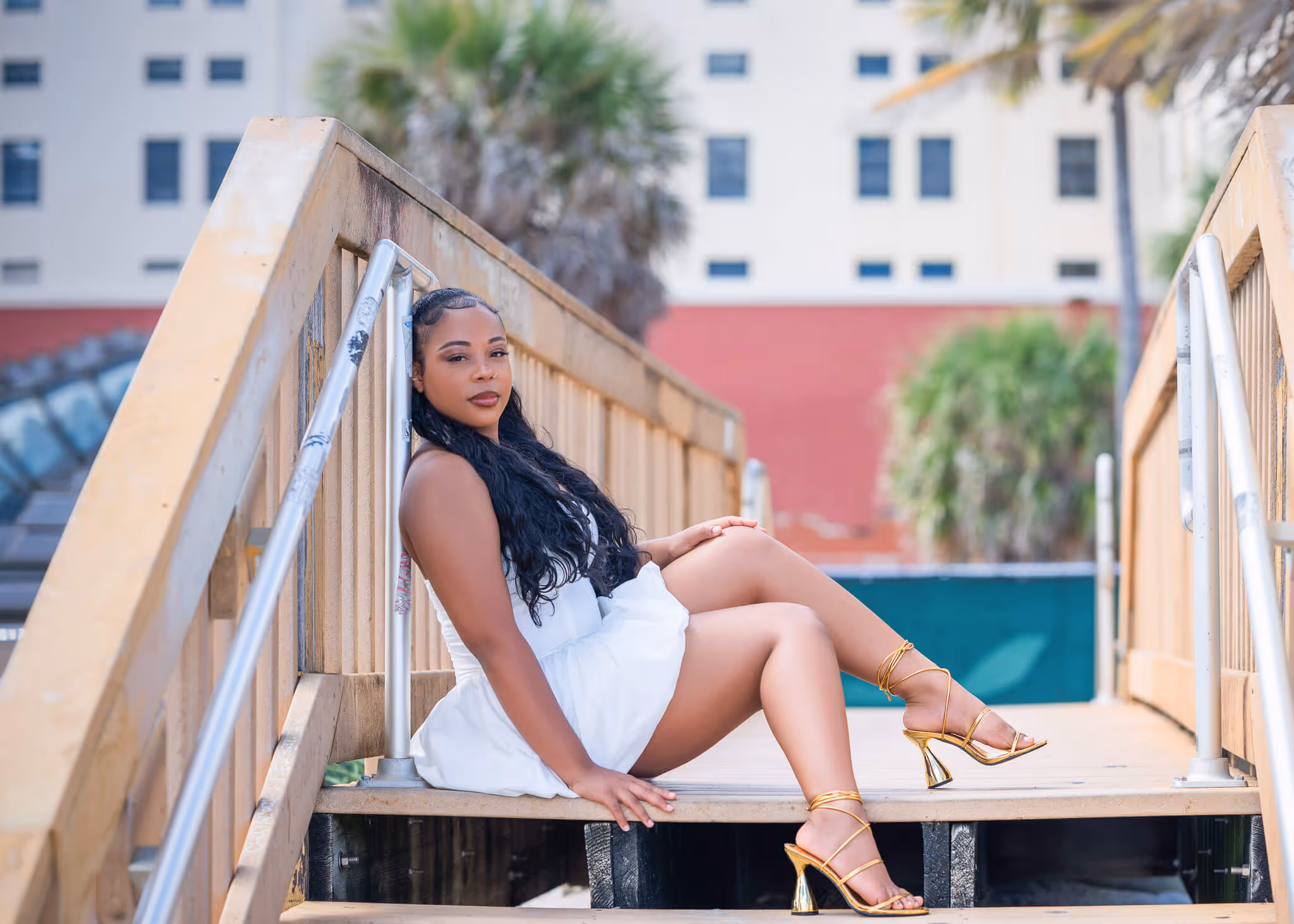 Woman with long black hair in a white dress and gold heels sitting on outdoor wooden stairs.