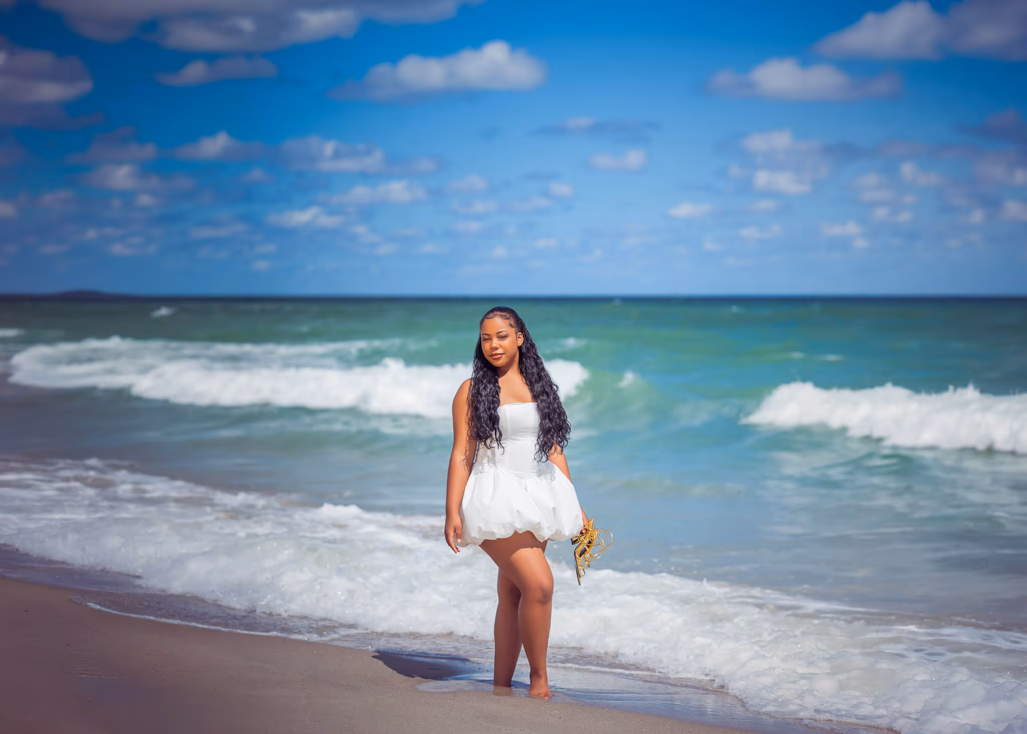 Woman in a short white dress standing barefoot at the edge of ocean waves on a sunny beach.