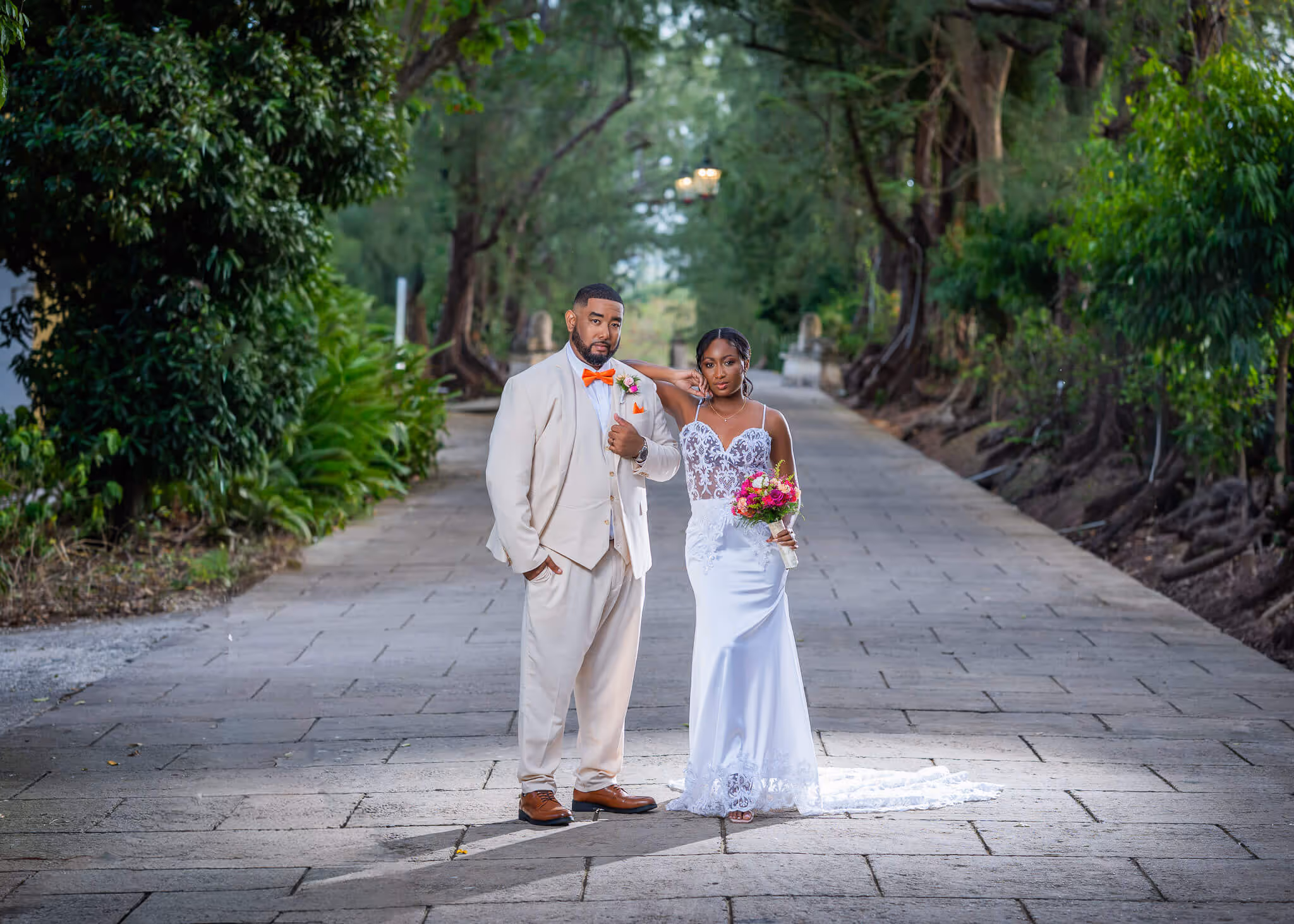 Bride in a white lace wedding dress holding a colorful bouquet standing next to groom in a beige suit with an orange bow tie on a tree-lined stone path.