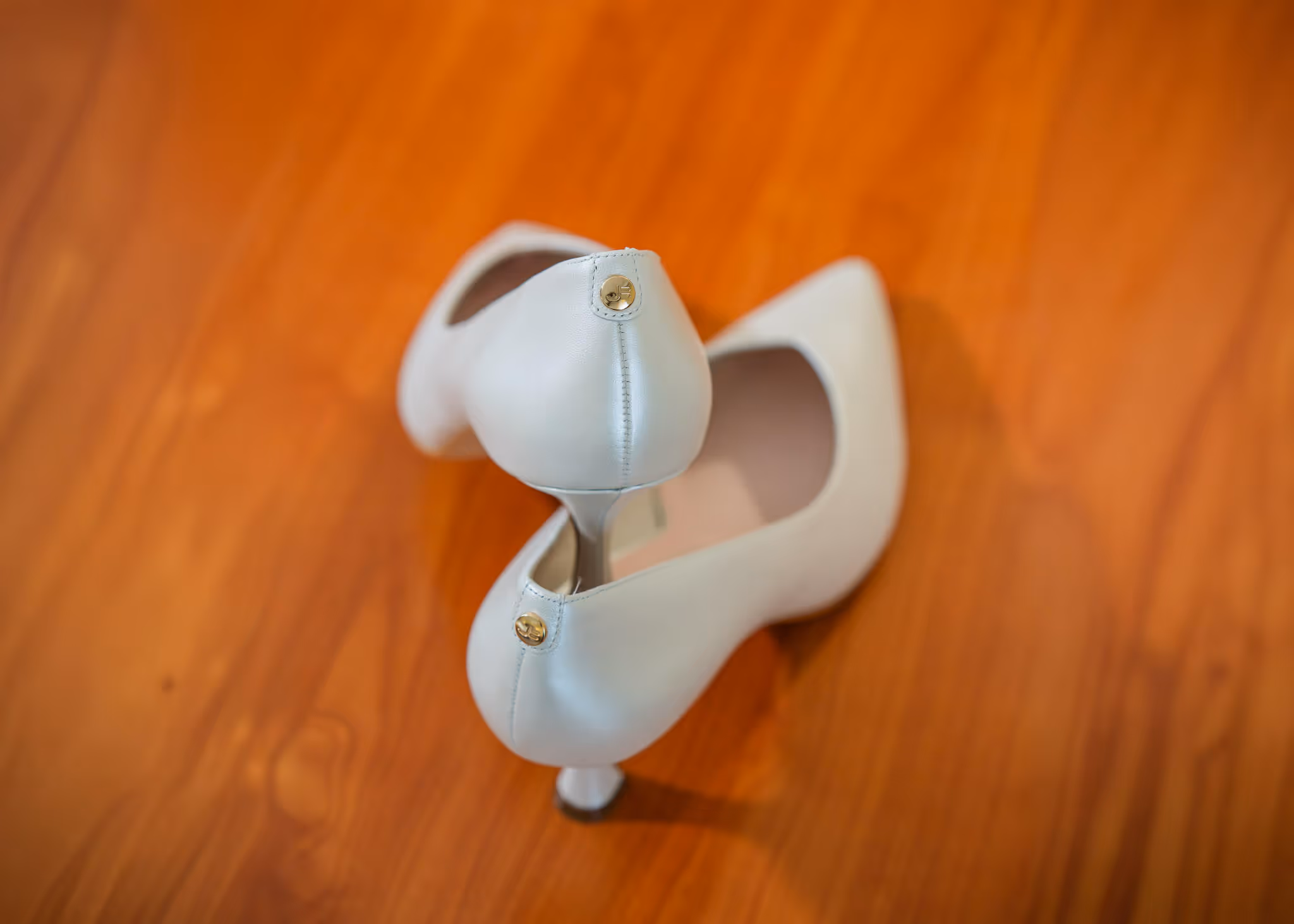 Pair of white high-heeled shoes with small gold buttons on the back, placed on a wooden floor.