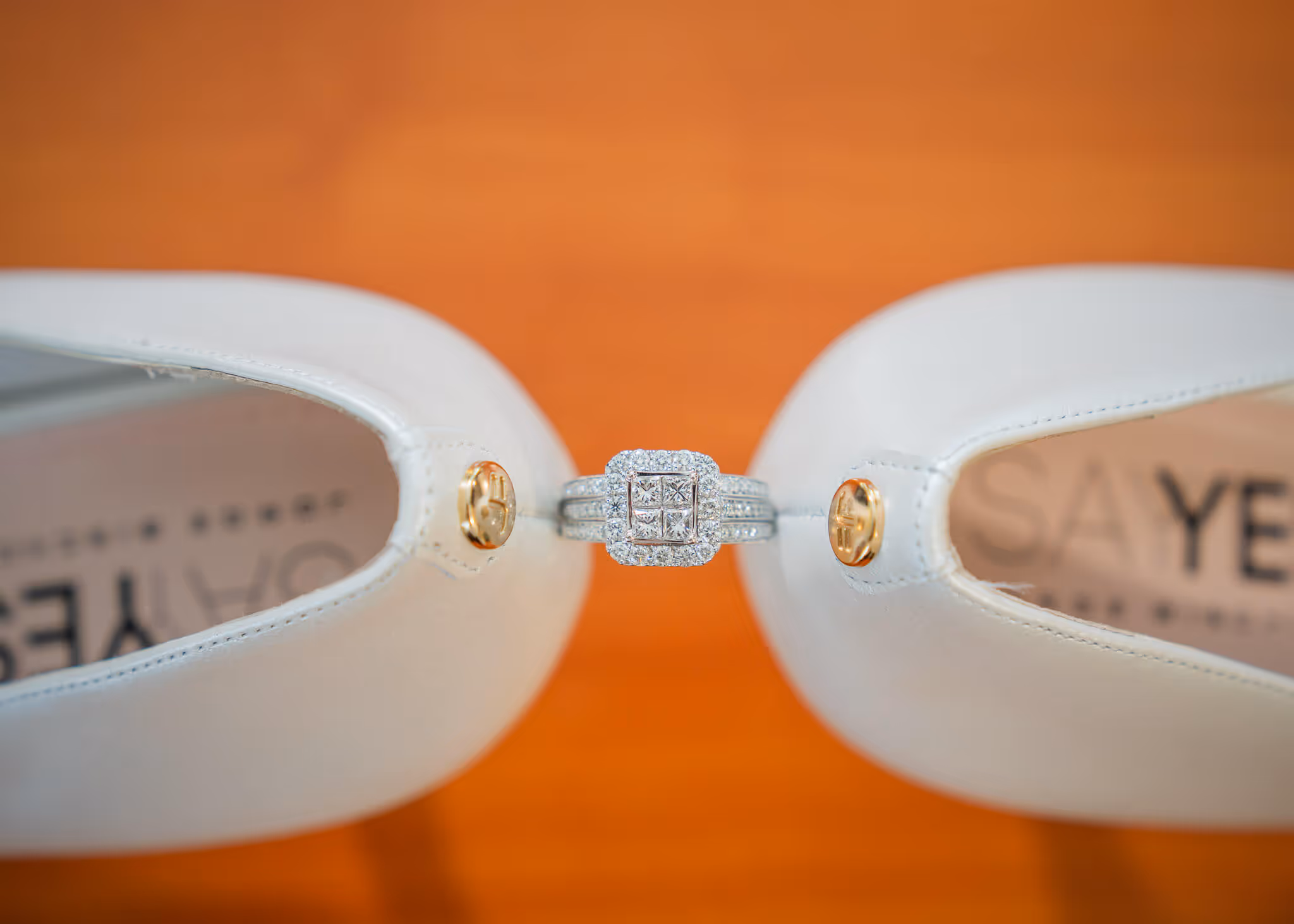 Diamond engagement ring placed between the heels of two white shoes on a wooden surface.