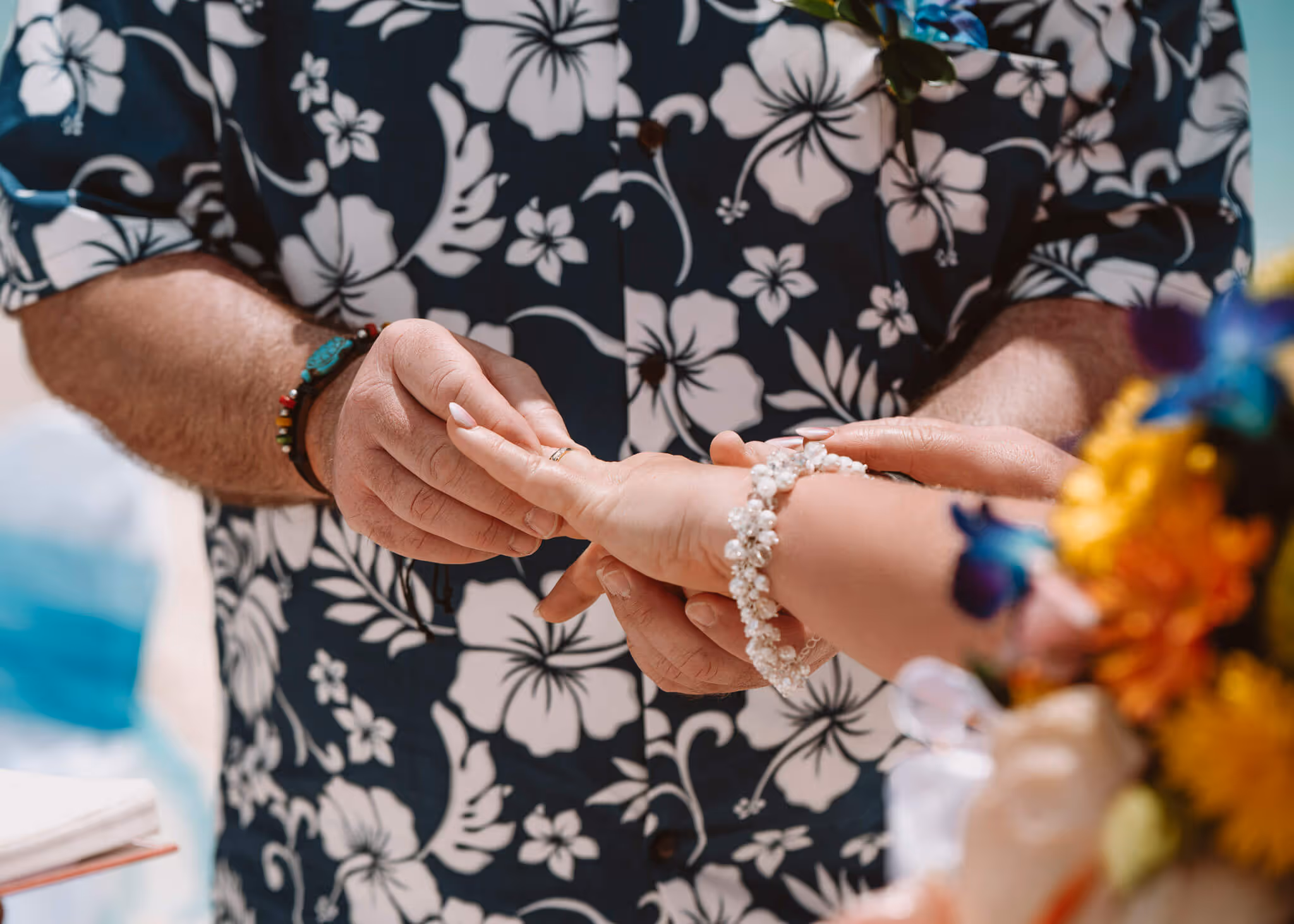Close-up of a person wearing a floral shirt placing a ring on another person's finger during a wedding ceremony.