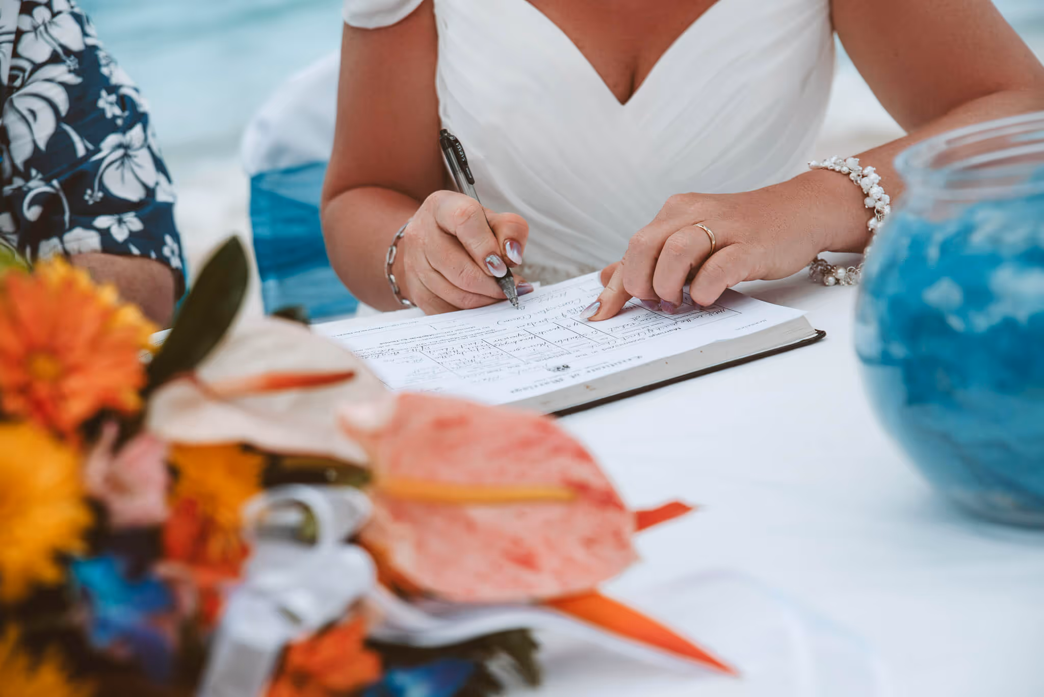 Bride in white dress signing a document on a table with colorful flowers and a blue decorative jar nearby.