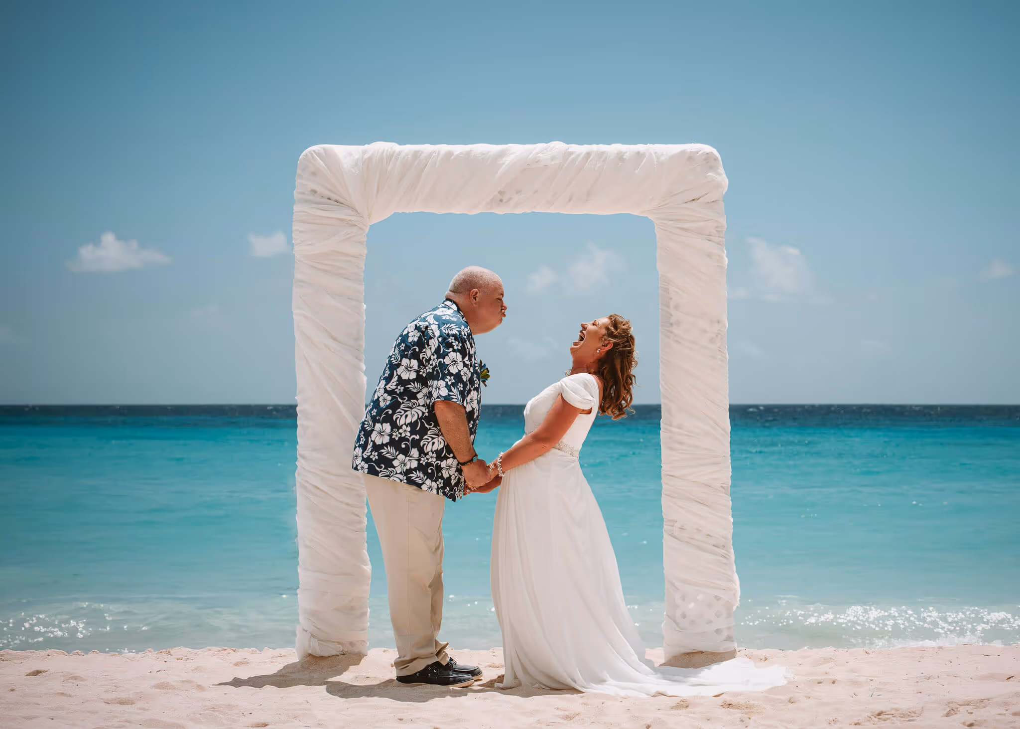 Couple holding hands and laughing under a white fabric arch on a sandy beach with turquoise ocean in the background.