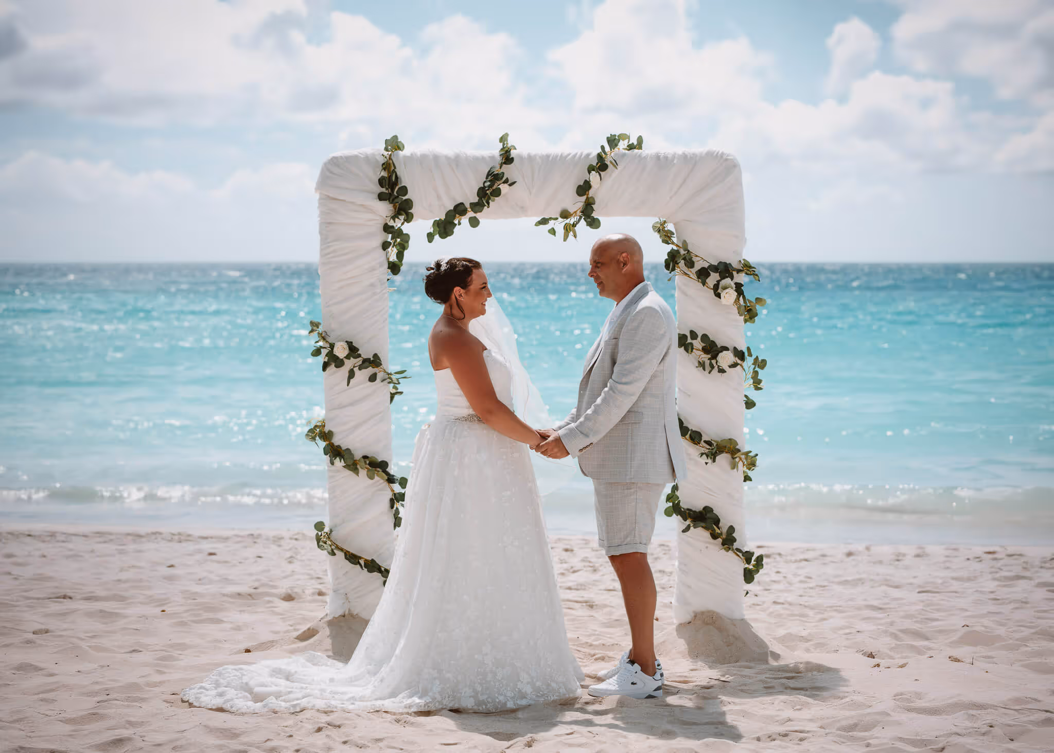 Bride and groom holding hands under a white decorated arch on a sandy beach with turquoise ocean in the background.