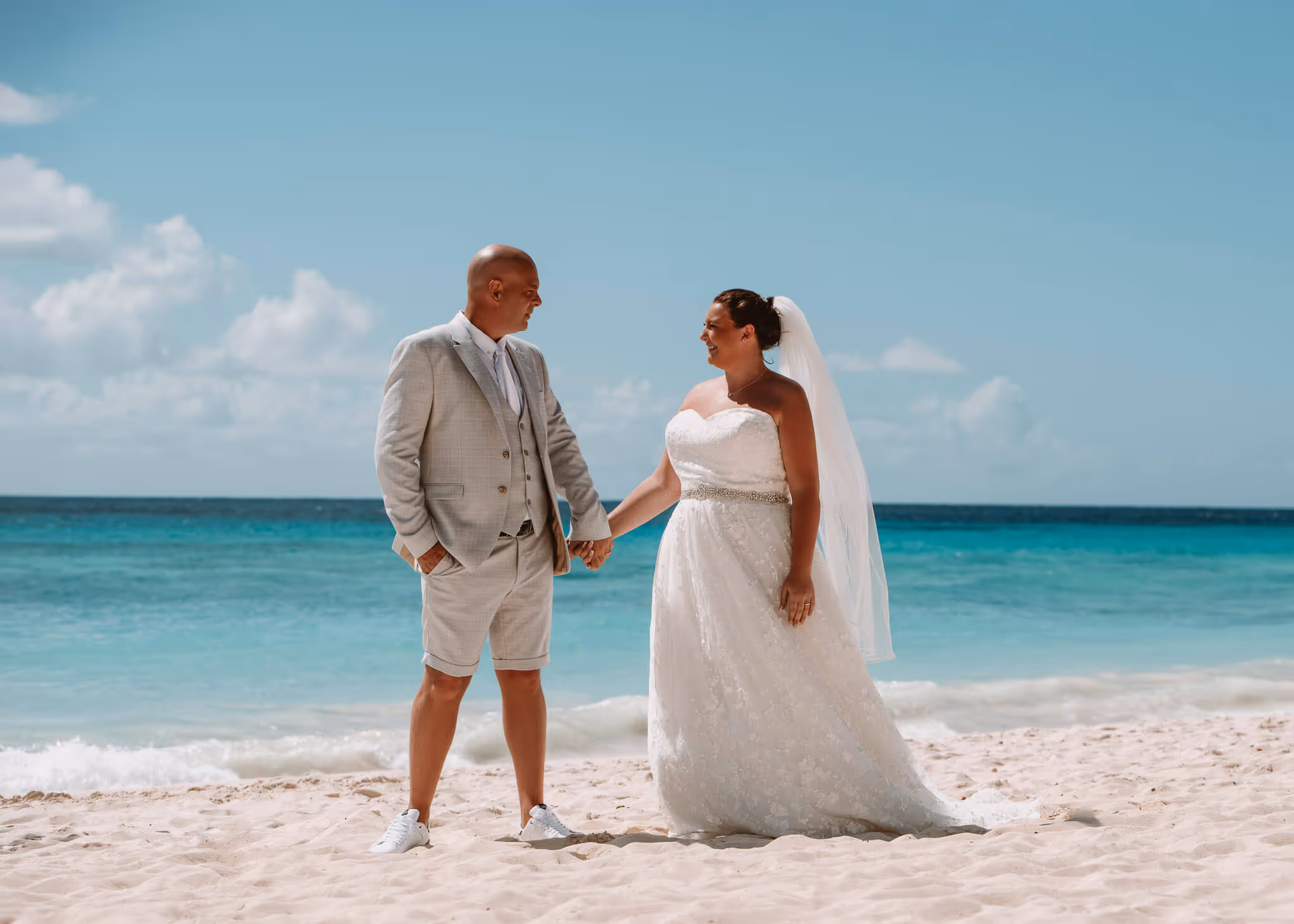 Bride in white wedding dress and veil holding hands with groom in light suit and shorts on sandy beach with ocean background.
