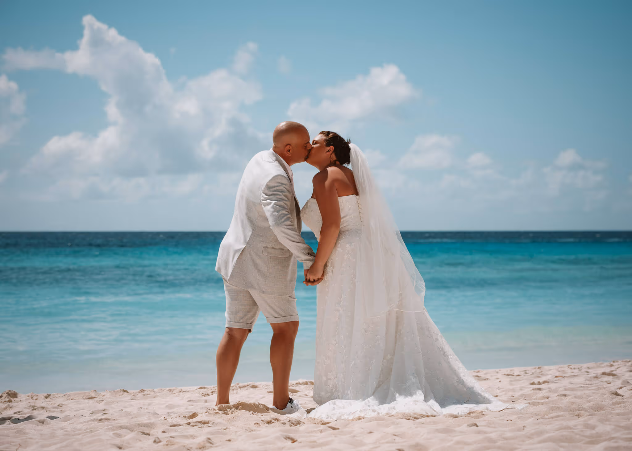 Bride and groom kissing on a sandy beach with turquoise ocean and partly cloudy sky in the background.